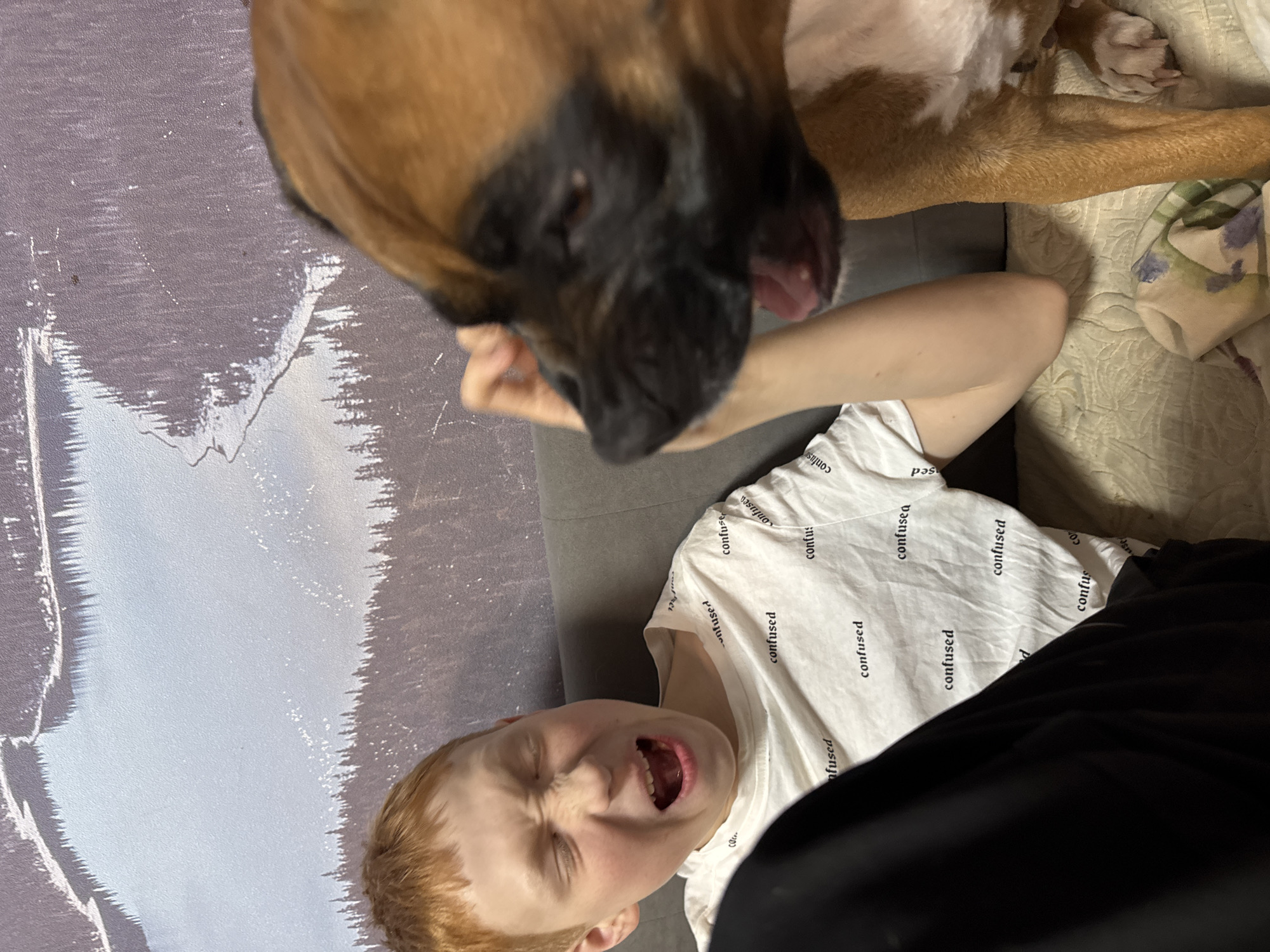 A boxer dog leans over a young red-haired boy lying on a couch wearing a white t-shirt. The boy is looking upward, his eyes closed and mouth open. The couch is gray, and a blanket is visible behind the boy. A wall with a pattern is visible to the left. The exact location of Chaikovsky, Russia, cannot be determined from this image.