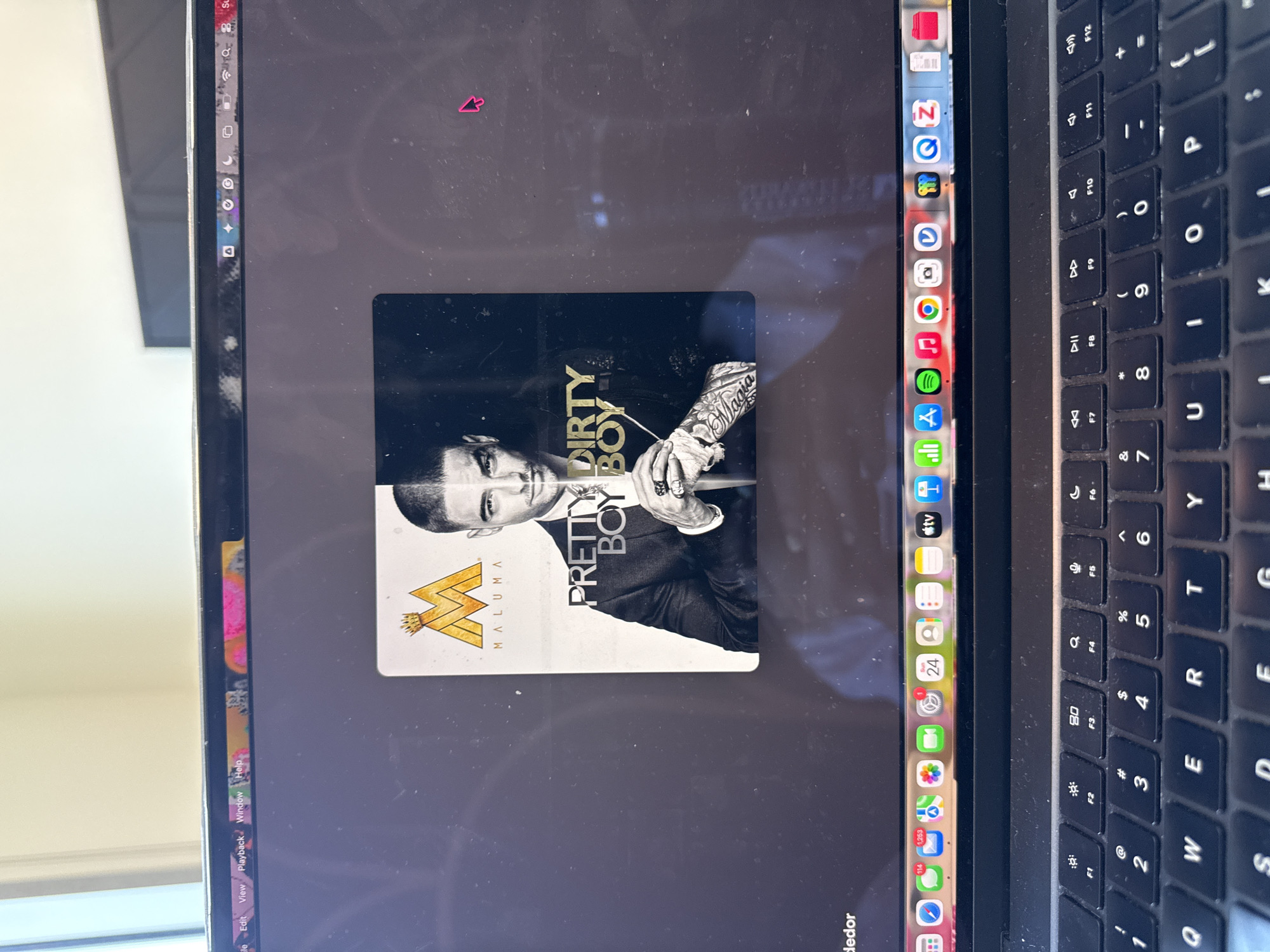 The image shows a laptop screen displaying an album cover with the title "Pretty Boy Dirty Boy" by Maluma. The laptop's keyboard and part of the screen's left-side menu bar are visible. The location within Austin, USA cannot be determined from the image.