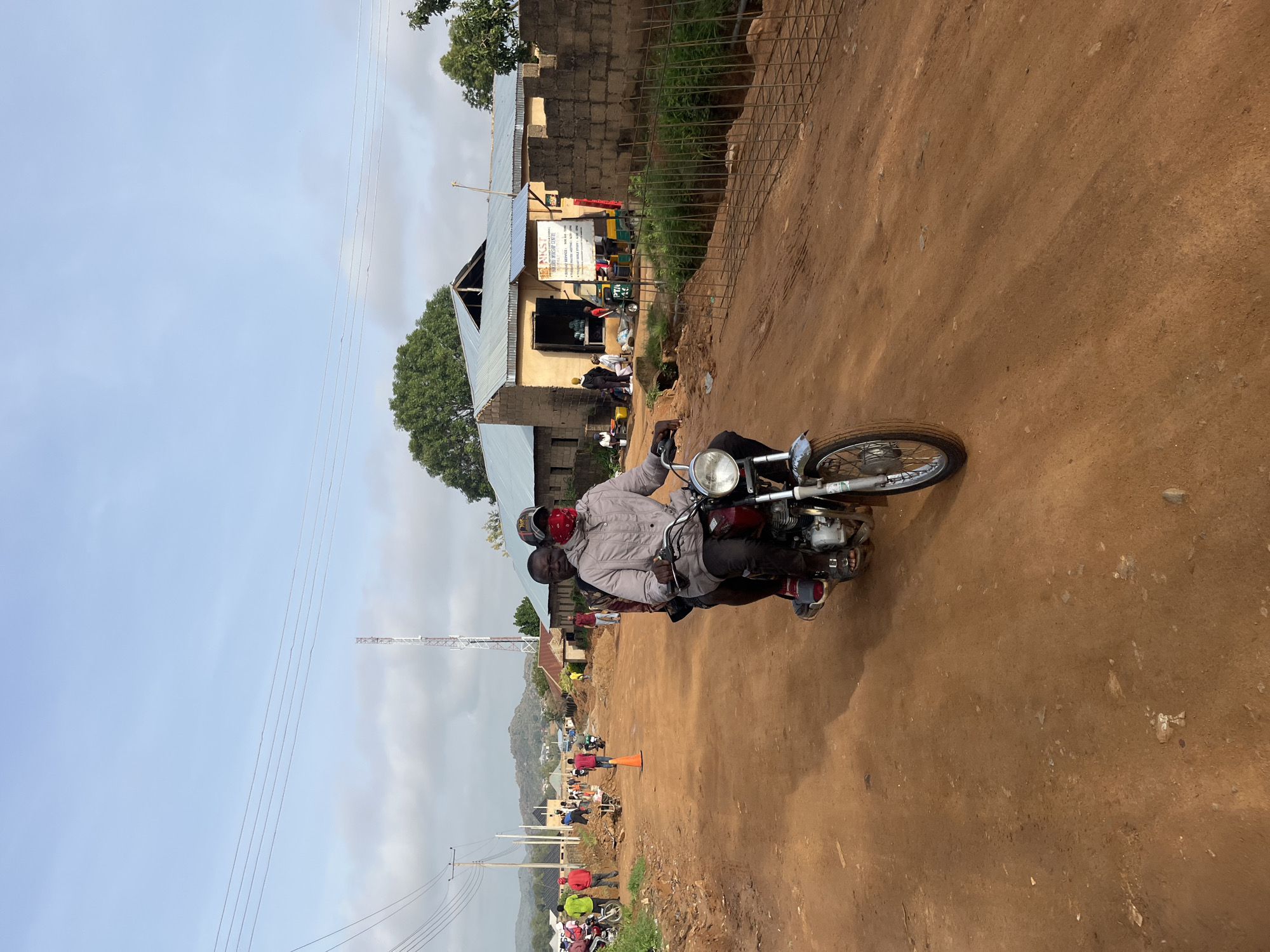 Two men are riding a motorcycle on a dirt road in Bukuru, Nigeria. The man in front is wearing a light-colored jacket and a red bandana over his face, and he is wearing a baseball cap. The man behind him is wearing a maroon shirt. The motorcycle is red and silver. The road is unpaved and dusty, with piles of dirt and construction materials on the sides. In the background, there are several buildings, including a church with a metal roof. There are also power lines and a cell tower. Several other people and motorcycles are visible in the background. The sky is partly cloudy.