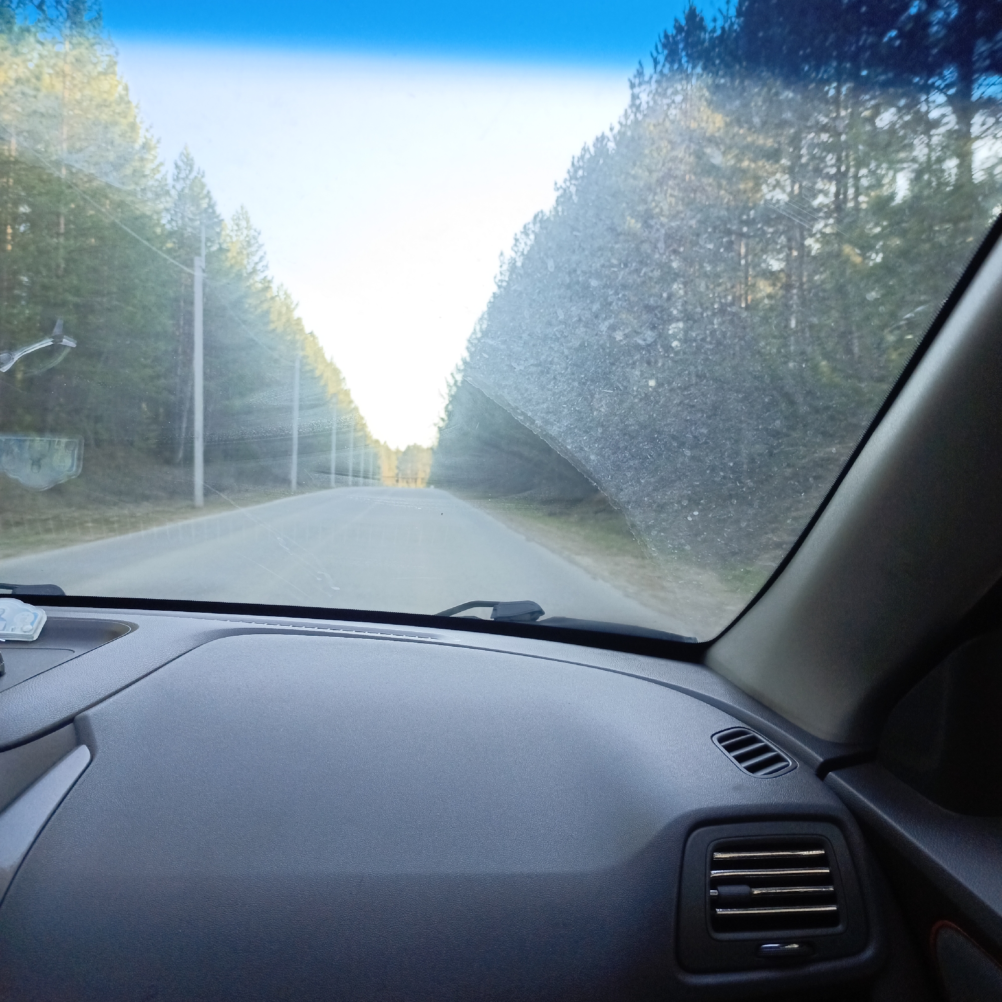 The image depicts a view from inside a car driving on a straight road, likely in a rural or forested area. The road stretches into the distance between rows of trees on both sides, with utility poles visible alongside the road. The car's dashboard is prominent in the foreground, showing air vents and other interior features. The windshield is partially obstructed by some dirt or condensation. The sky is visible above the trees, appearing bright.