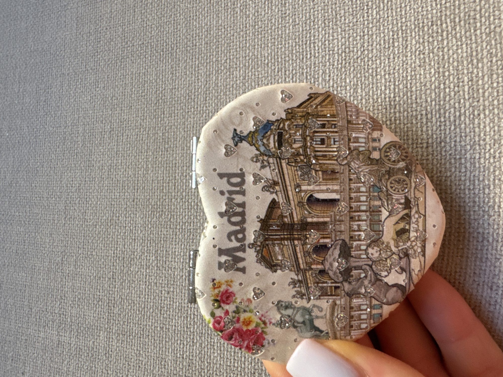 A heart-shaped compact mirror is held in a person's hand against a textured gray background. The compact is decorated with an illustration of a cityscape, the word "Madrid" in large silver lettering, and various embellishments. The illustration depicts a grand building with arched entrances and domes, a statue of a person on a horse, a bear, and a lion pulling a chariot. Small, sparkling heart-shaped rhinestones are scattered across the surface of the compact. The background appears to be a coarse fabric, possibly a tablecloth or carpet. There is no indication of the city of Murcia, Spain in this image.