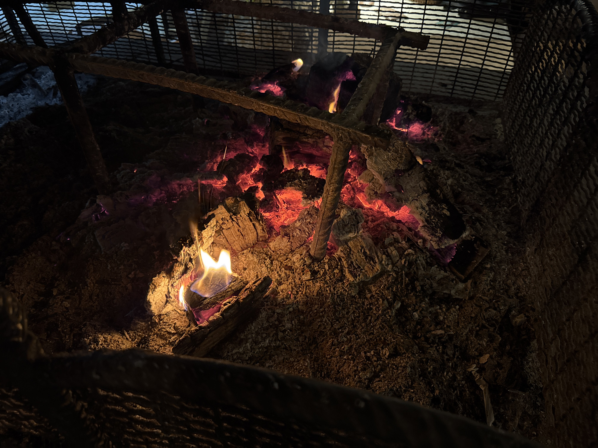 A fire pit is viewed from above. Embers glow red and orange amidst dark ash and partially burned wood. Flames flicker around the wood. A metal frame surrounds the fire. Overhead, a metal grid suggests the fire pit is located outdoors. No people are visible. The location of Podgorica, Montenegro is irrelevant to the observable content of the image.