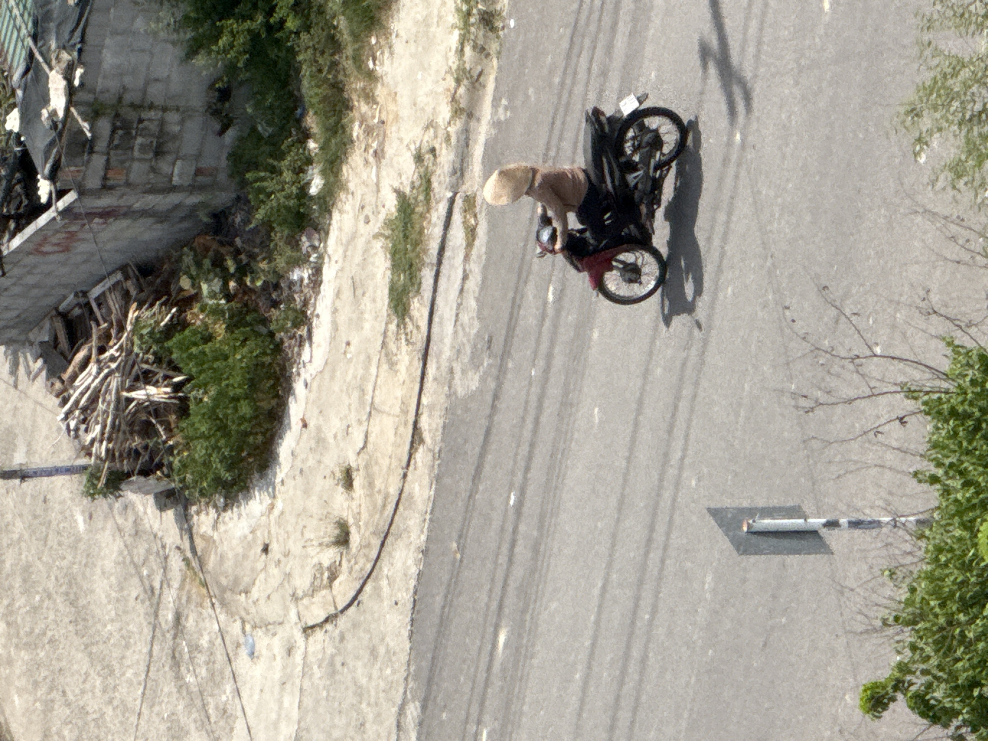 A woman wearing a conical hat and a brown shirt rides a red and black motorcycle on a paved road in Bac Nha Trang Ward, Vietnam. She is facing right and appears to be moving forward. The road is gray with visible tire tracks and scattered white debris. To the left of the road, there is a patch of green foliage and a gray signpost with black and white stripes. In the background, there is a light-colored, rough terrain area with scattered green vegetation and a partially constructed building with a gray, weathered wall and a black roof. Several dark power lines are visible spanning across the upper portion of the image.
