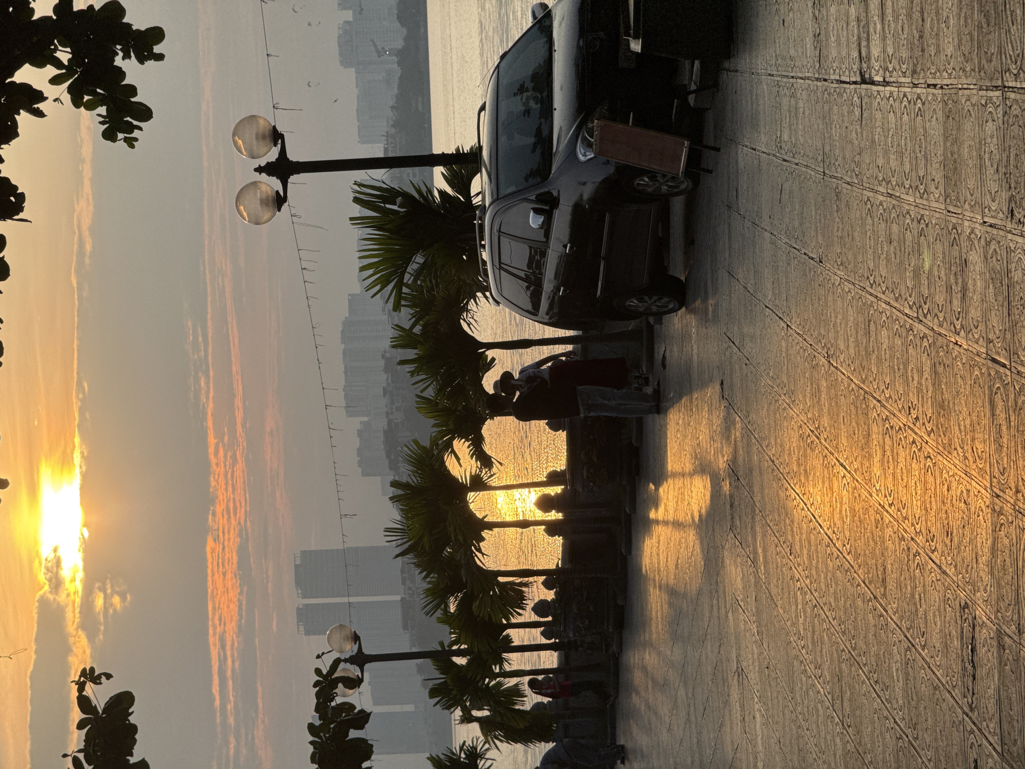 This is a vertical, eye-level, medium shot taken at sunset on the west side of West Lake in Hanoi, Vietnam. A dark-colored minivan is parked on a paved walkway in the foreground, facing left and angled away from the camera. A person wearing a hat and a long skirt stands near the front of the minivan, also facing left and away from the camera. A couple stands in silhouette further to the left, with their arms around each other. They are standing in front of a low stone wall behind which a line of palm trees is visible. The water of West Lake is to the right of the wall, and the sunset is reflecting on the surface. The sky is filled with orange and yellow clouds. In the background, hazy outlines of apartment buildings can be seen. The paved walkway in the foreground is illuminated by the setting sun, creating long shadows.