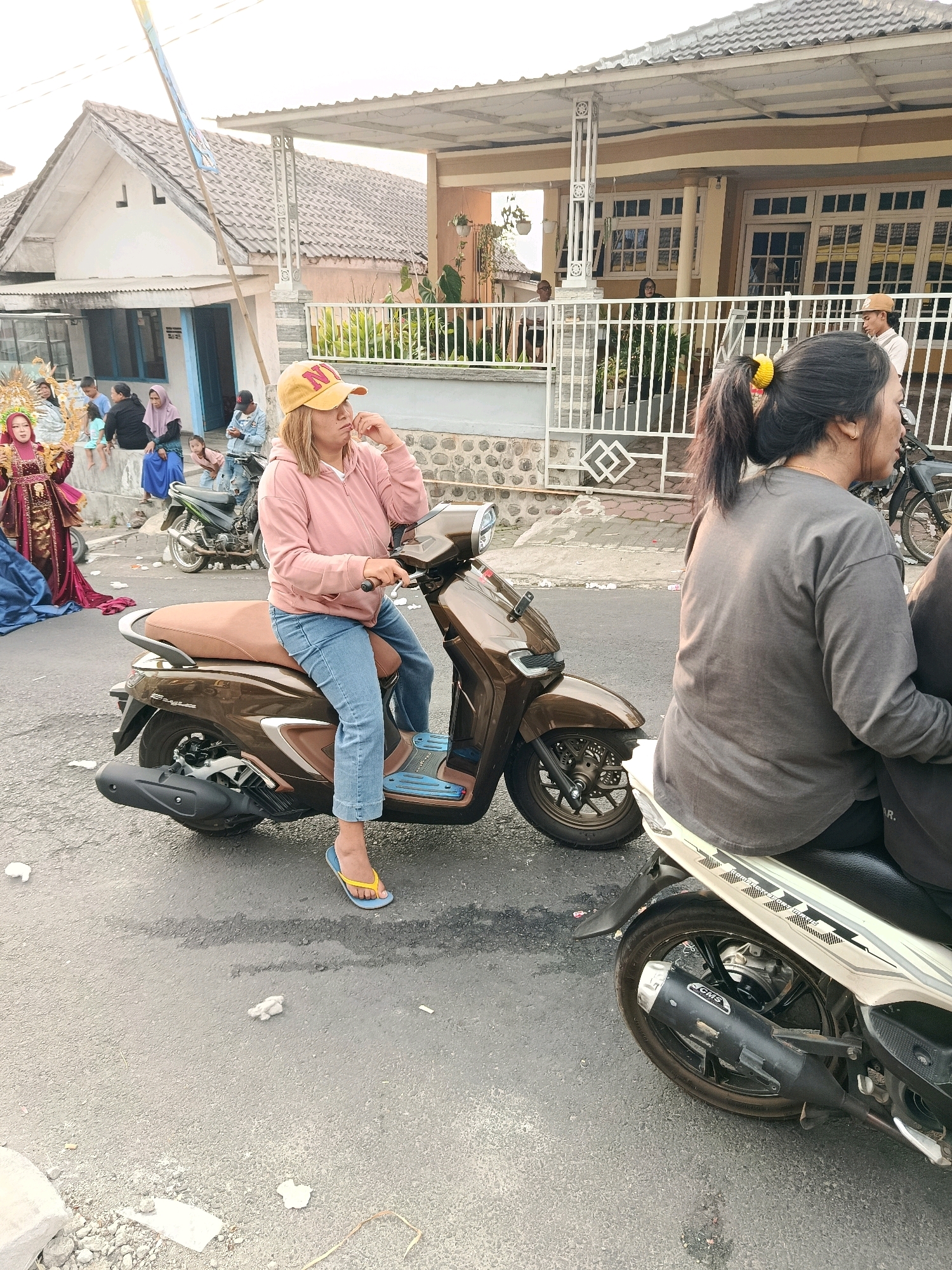 The image depicts a street scene in Gubukklakah, Indonesia, with several people and motorcycles. In the foreground, a woman wearing a pink hoodie, blue jeans, and a yellow baseball cap is seated on a brown scooter. She is looking to the right and touching her chin with her hand. To her right, another person, partially visible, is seated on a white scooter, also facing away from the camera. In the background, a group of people are gathered, including a person in a traditional red and gold outfit. Several houses and a white fence are visible, suggesting a residential area. The ground is paved asphalt, with some debris scattered on it. The lighting indicates it is daytime, possibly late afternoon or early morning given the soft light.