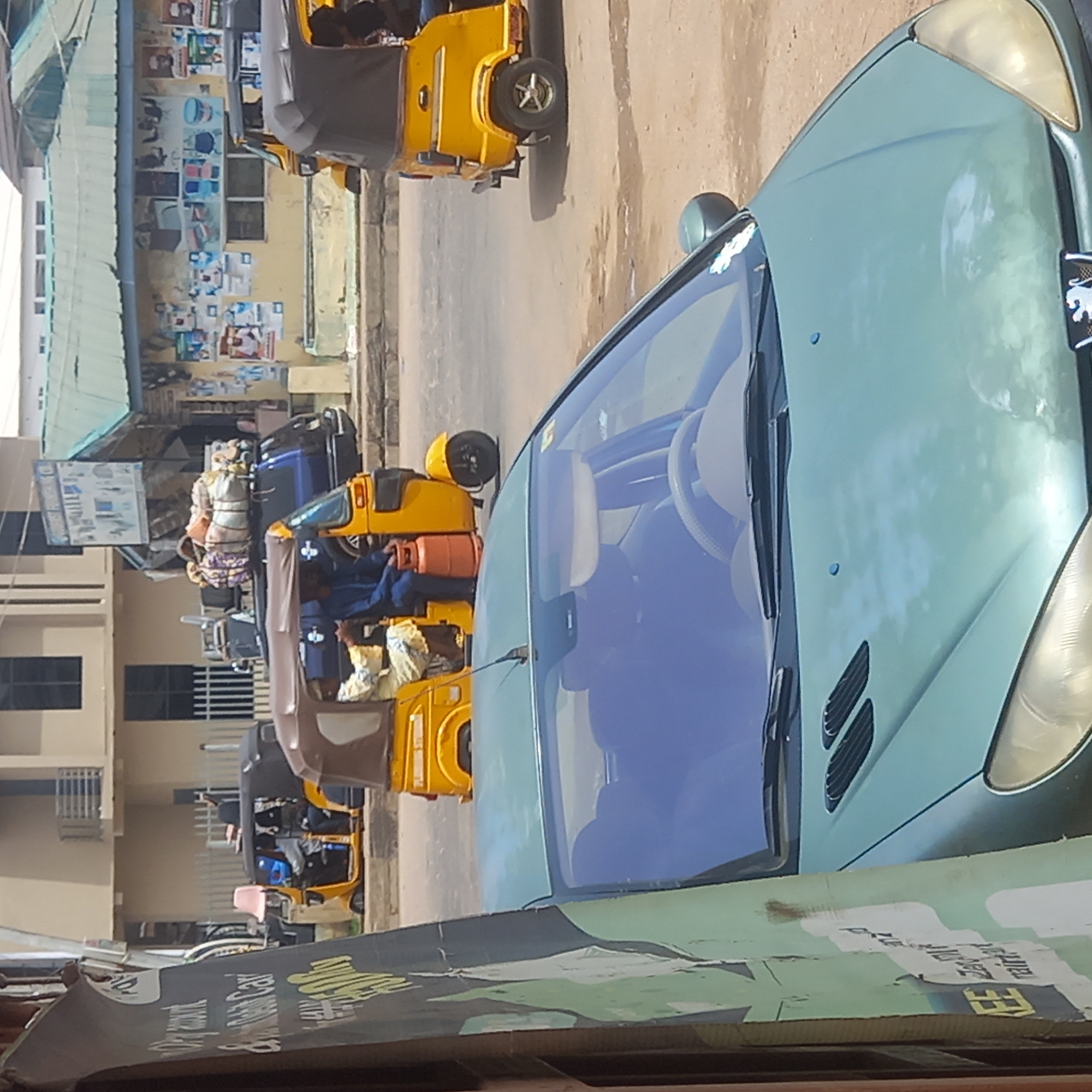 The image depicts a street scene in Dadi Riba, Nigeria. In the foreground, there's a section of a car, specifically its hood and windshield, with the interior visible. Behind the car, the street contains several yellow auto-rickshaws, some of which appear to be carrying passengers or cargo. A building lines the edge of the street, its walls covered with posters or advertisements. The overall scene presents a busy urban environment.