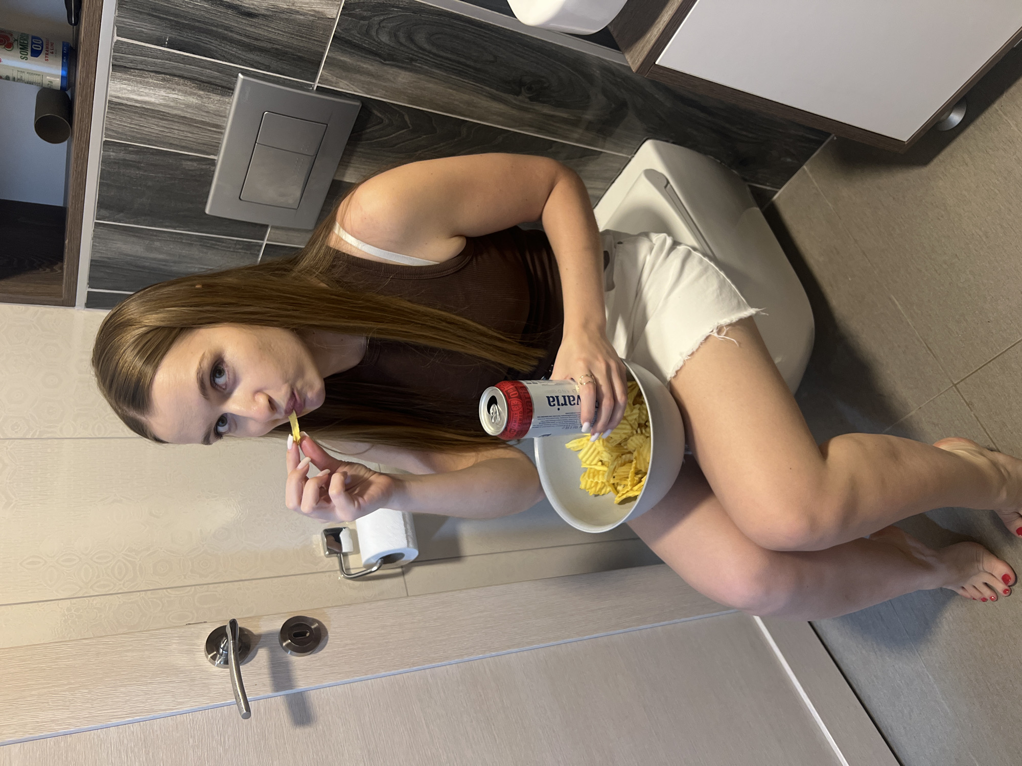 A young woman is seated on a toilet in a bathroom. She wears a brown tank top and white shorts. She holds a bowl of chips and a can of drink labeled "Varia". She is in the act of eating a chip. The bathroom has grey and white tiles, a toilet paper holder, a toilet flush button, a door with a handle, and a cabinet above the toilet. There is no information about the city Budva, Montenegro.