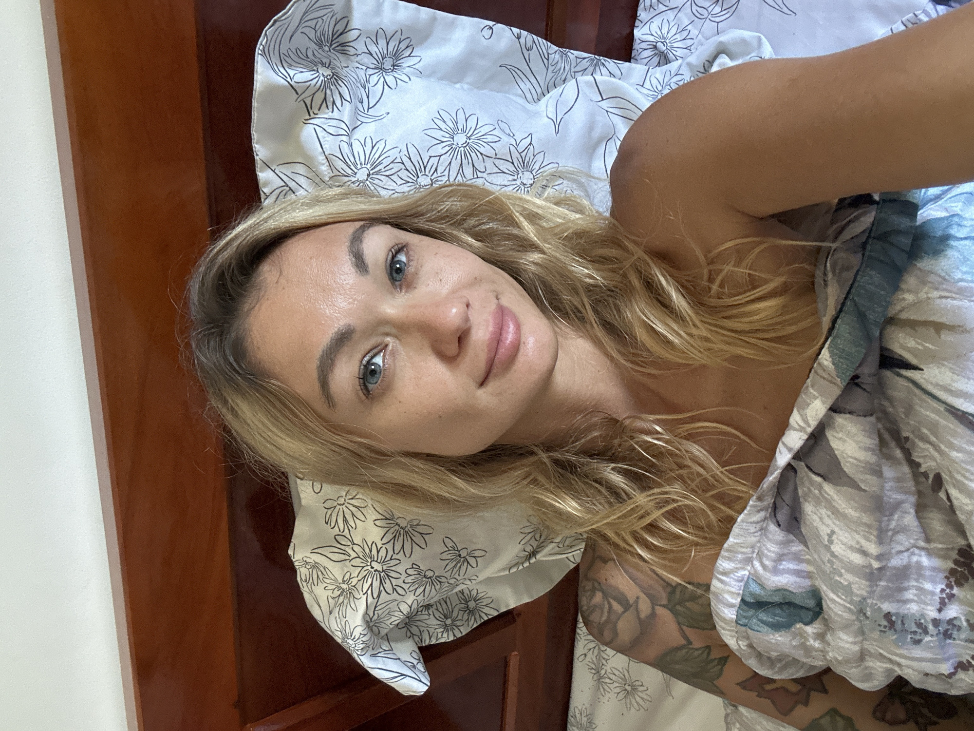 A young Caucasian woman with blonde hair and blue eyes is lying in a bed. She is looking at the camera and smiling slightly. Her hair is long and wavy, and it falls around her shoulders. She is wearing a strapless top and a blanket is pulled up to her chest. There is a tattoo on her left arm and shoulder. The bed is made with white sheets and a duvet with a floral pattern. The headboard of the bed is made of dark wood. The background is blurred, but it appears to be a room with white walls. The lighting is natural, coming from the left side of the image. The photo was taken in Mũi Né, Vietnam.