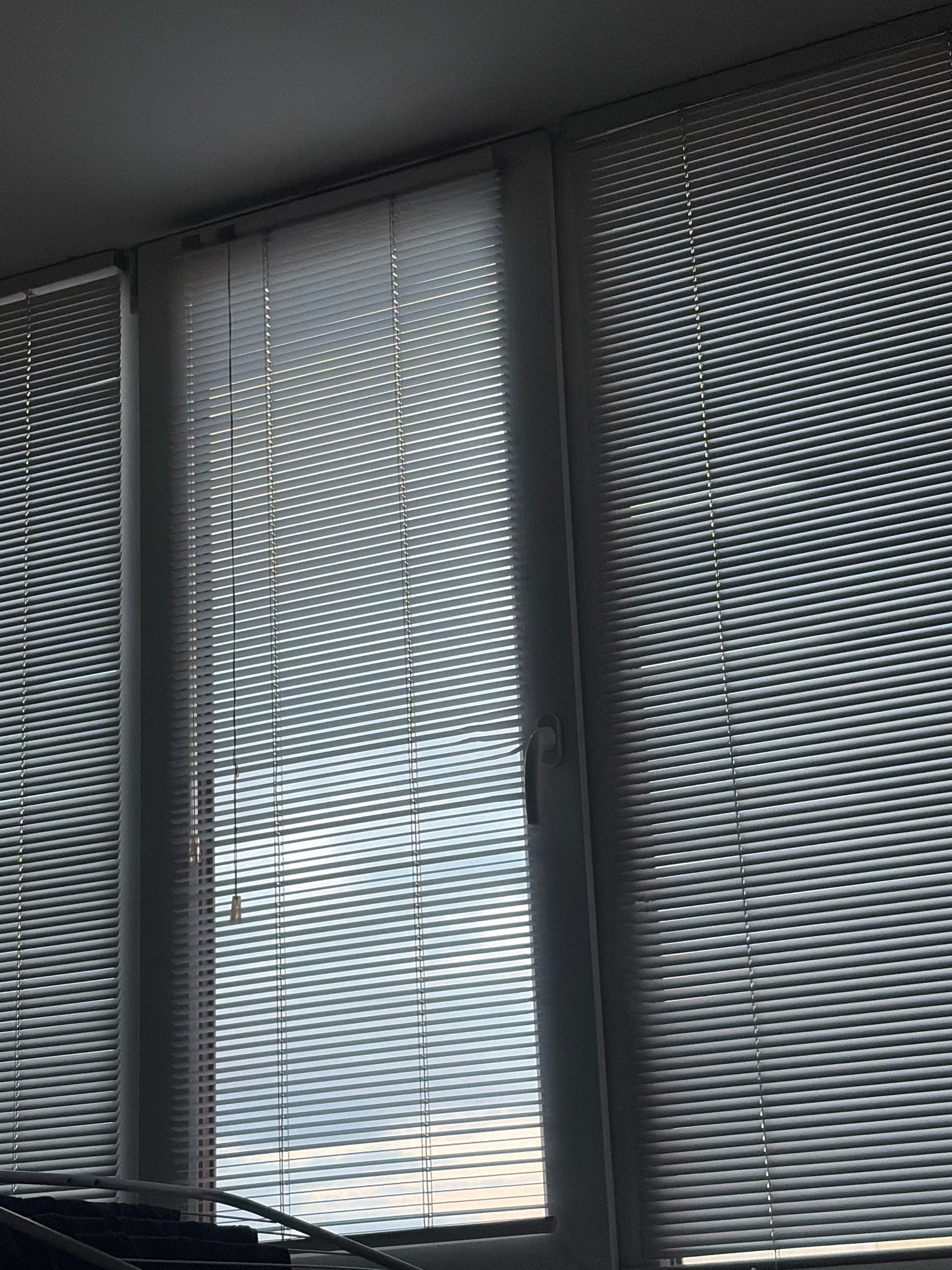 The image displays a window covered by white horizontal blinds. The blinds on the left and right are closed, while the blinds in the center appear to be partially open, allowing a view of a bright sky with some clouds. A white cord for adjusting the blinds hangs down in the center. A window handle is visible on the right-hand side of the central window. In the lower portion of the image, a dark object, possibly a piece of furniture or clothing, is partially visible. The scene is indoors, suggesting an apartment or office setting. There is no information in the image to suggest a specific location in Sholomovo, Russia.