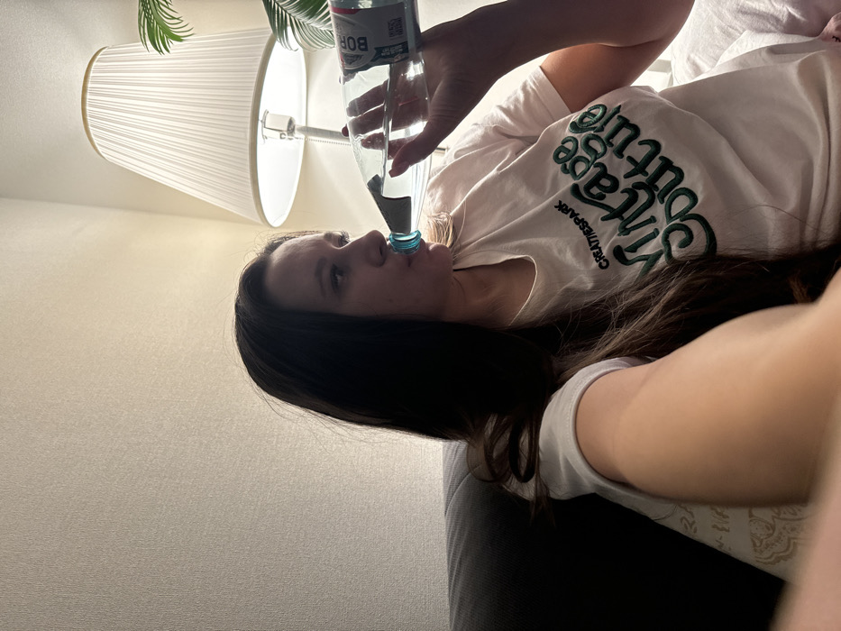 A woman is lying down, angled to the left, drinking from a clear plastic water bottle labeled "BOR". She wears a white t-shirt with "Village Couture" embroidered in green. Her dark hair cascades over her shoulder. In the background, a white wall meets a lamp with a pleated shade and a potted plant. The image appears to be a selfie taken at a close range, with a focus on the woman's face and the water bottle.