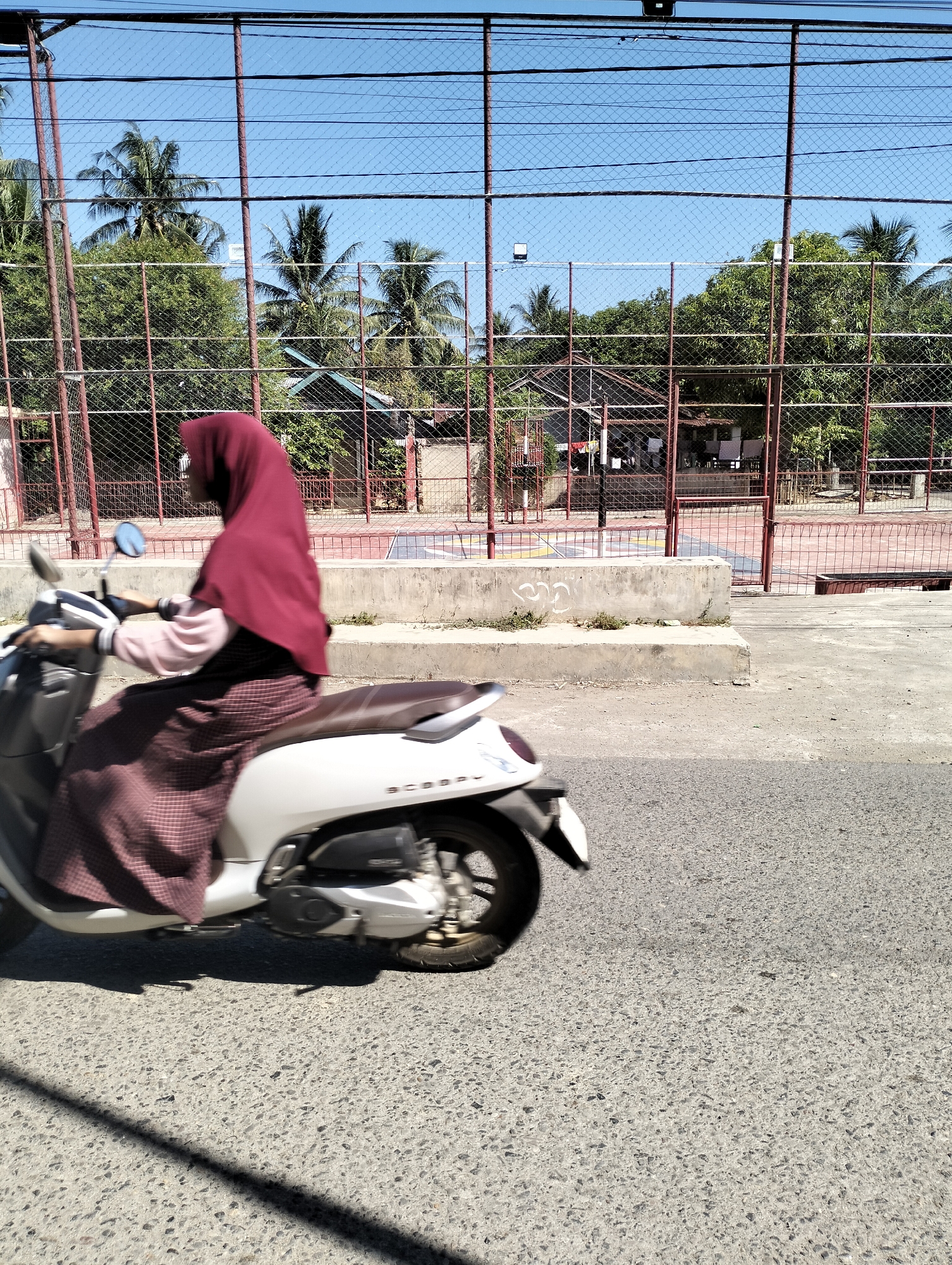 A woman wearing a burgundy hijab and a patterned skirt rides a white scooter on a paved road. The scooter has a brown seat and the word "SCOOPY" is visible on its side. In the background, a fenced-in sports court with a red surface and white markings is visible. Trees, including palm trees, and a few buildings are also present behind the court, under a clear blue sky. The image appears to be taken during the daytime. The specific location mentioned, "Janggotseungko, Indonesia," is not explicitly identifiable from the visual elements in the image.