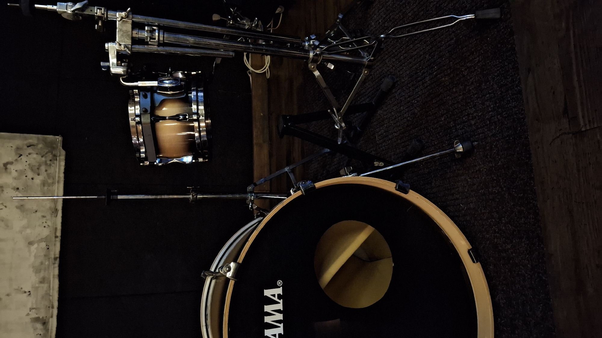 A vertical, medium shot captures a drum set on a dark carpeted floor in an indoor setting. In the foreground on the left is a large, black bass drum with a visible "TAMA" logo. Its wooden rim is partially visible, and the inside of the drum is a golden color. To the right of the bass drum, a smaller, tan-colored tom-tom drum is positioned on a stand. This drum has chrome hardware and a wooden shell with a gradient finish. Behind the tom-tom, a complex metal stand with multiple arms and joints, likely a cymbal stand, is visible. The background consists of a dark, textured wall, possibly acoustic paneling. Above the drums, a pale, mottled surface, possibly a banner or backdrop, is seen against the dark wall. The lighting is dim, casting shadows and highlighting the metallic components of the drum set. No people are present in the image.