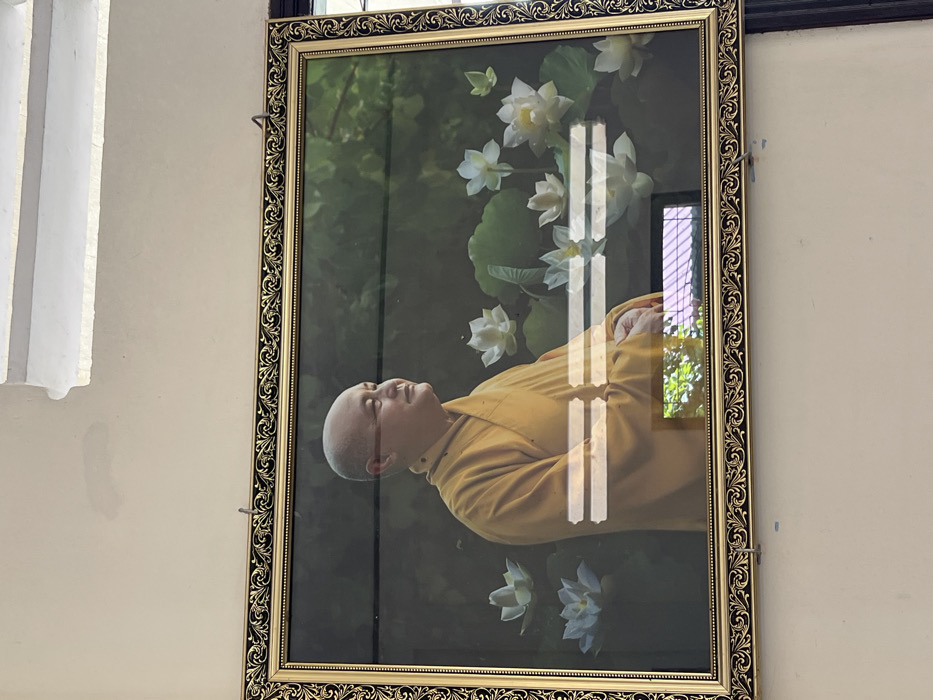 A framed portrait hangs on a beige wall. The subject appears to be a monk in yellow robes, lying down among white lotus flowers. The frame is ornate gold with a dark pattern. Reflections of a window and outdoor greenery are visible on the glass surface of the portrait. A white, architectural element is partially visible on the left side of the image. The wall also shows minor imperfections.
