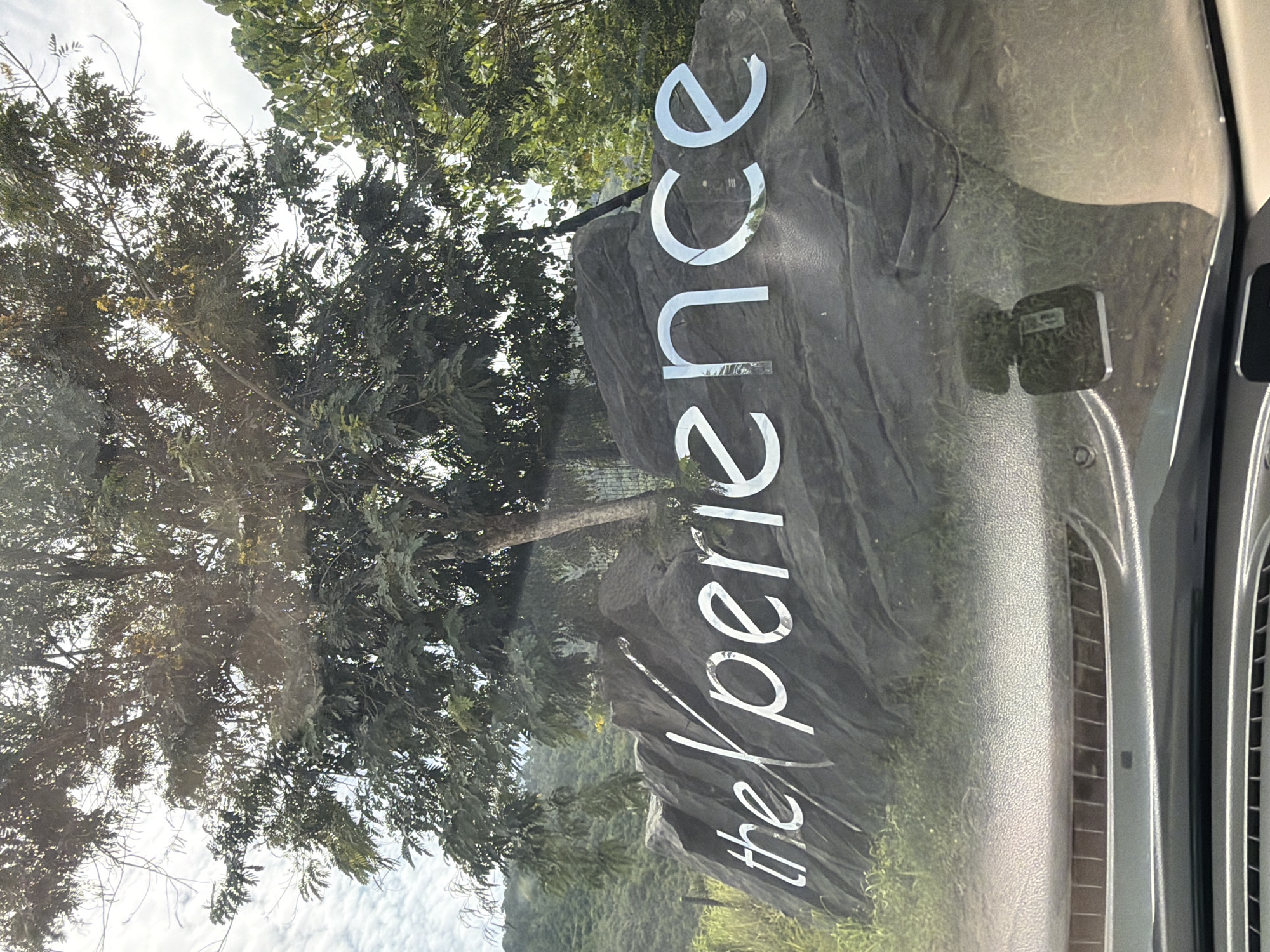 In the city of Ko Samui District, Thailand, a large rock formation is the backdrop for the metallic sign that reads "the Xperience." The letters are a bright silver, contrasting with the dark, textured surface of the rock. The sign is positioned in an outdoor setting, with a large tree in the foreground, its branches extending over the rock and sign. The leaves of the tree are dense, with sunlight filtering through them, creating a dappled effect. In the background, lush green hills rise, covered in vegetation. The sky is overcast with soft, white clouds. A car's dashboard and windshield are visible in the bottom portion of the image, indicating the photo was taken from inside a vehicle. The windshield reflects the environment, including the car's interior and some exterior elements.