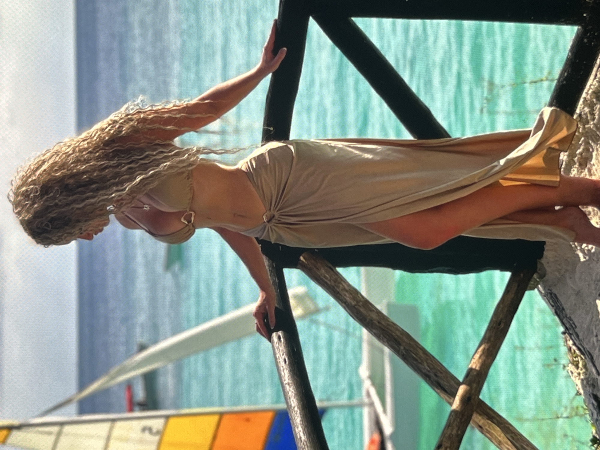 A woman with long, curly blonde hair stands on a wooden platform, leaning on a railing. She is wearing a tan bikini and a matching sarong with a high slit. Her body is partially visible from the side. In the background, there is a body of turquoise water with a sailboat and a distant red boat. The sky is overcast. The location is described as Unknown, Unknown.