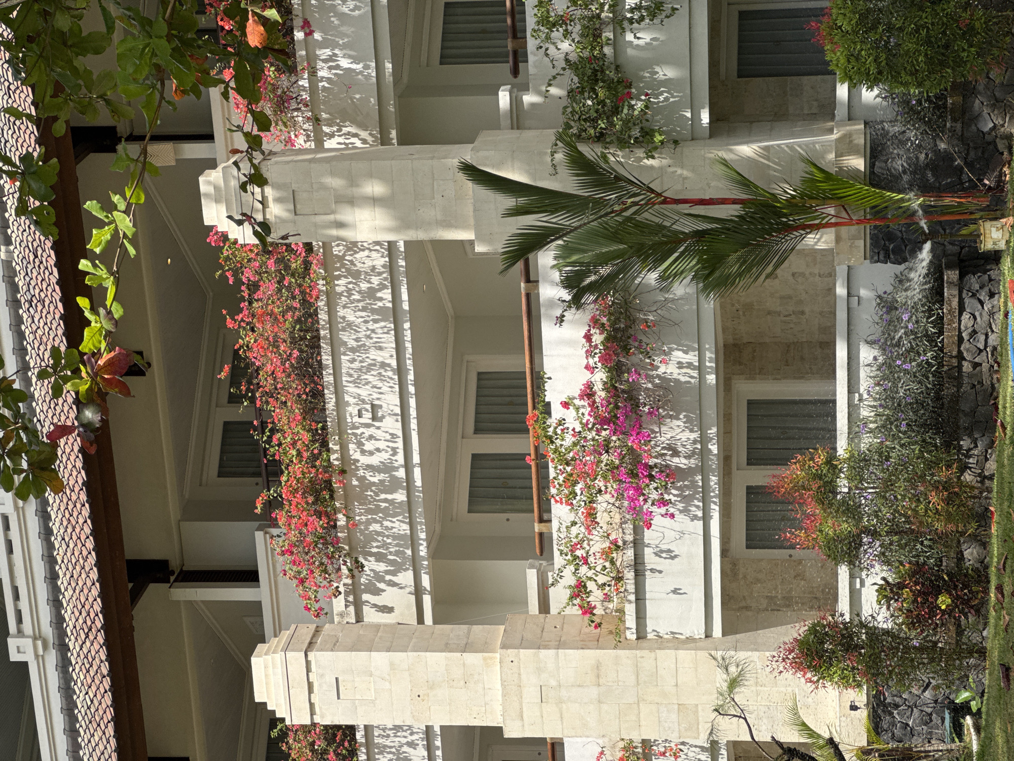 The image shows a multi-story building with balconies adorned with vibrant pink and red bougainvillea flowers. The building's facade is white, with stone accents on the pillars and lower walls. Palm trees and other lush green vegetation are visible in the foreground and on the balconies, suggesting a tropical environment. The lighting indicates it is daytime, with sunlight casting shadows on the building's exterior. The scene appears to be a resort or hotel in Kuta Selatan, Indonesia, characterized by its tropical flora and architecture. There are no people visible in the image. The atmosphere is serene and picturesque, highlighting the natural beauty and architectural style of the location.