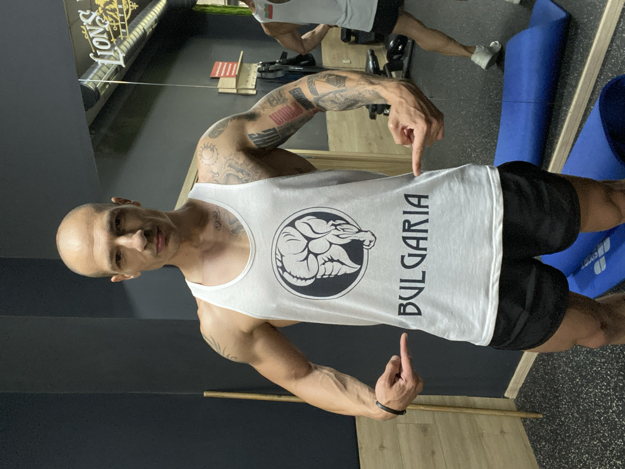 A man is in a gym with a mirror. He is wearing a white tank top with a logo containing a lion and the text "BULGARIA". He is wearing black shorts. He has tattoos on his arms. He is pointing to the logo on his shirt and giving the middle finger. In the mirror, another person is seen lifting weights, wearing a white tank top. There is a blue mat in the background near the mirror. A sign reading "lions fit" is visible in the background. The location is Pleven, Bulgaria.