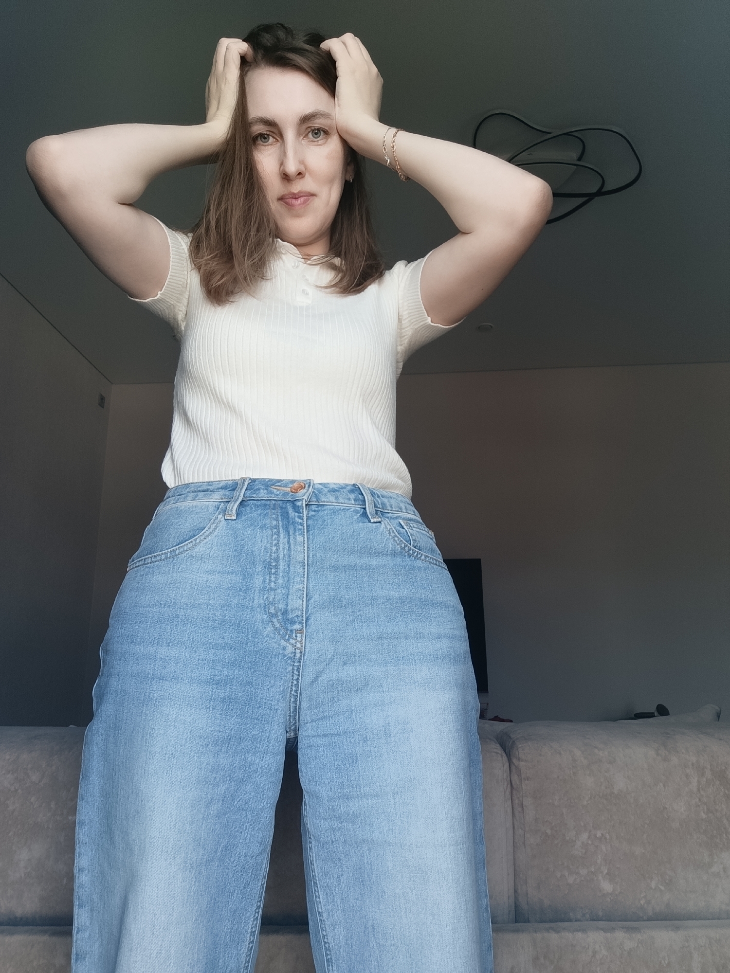 A woman stands indoors, facing the camera, with her hands on her head. She wears a cream-colored ribbed top and light blue wide-leg jeans. A couch is visible in the background, along with a television and a modern light fixture on the ceiling. The location is Chaikovsky, Russia.