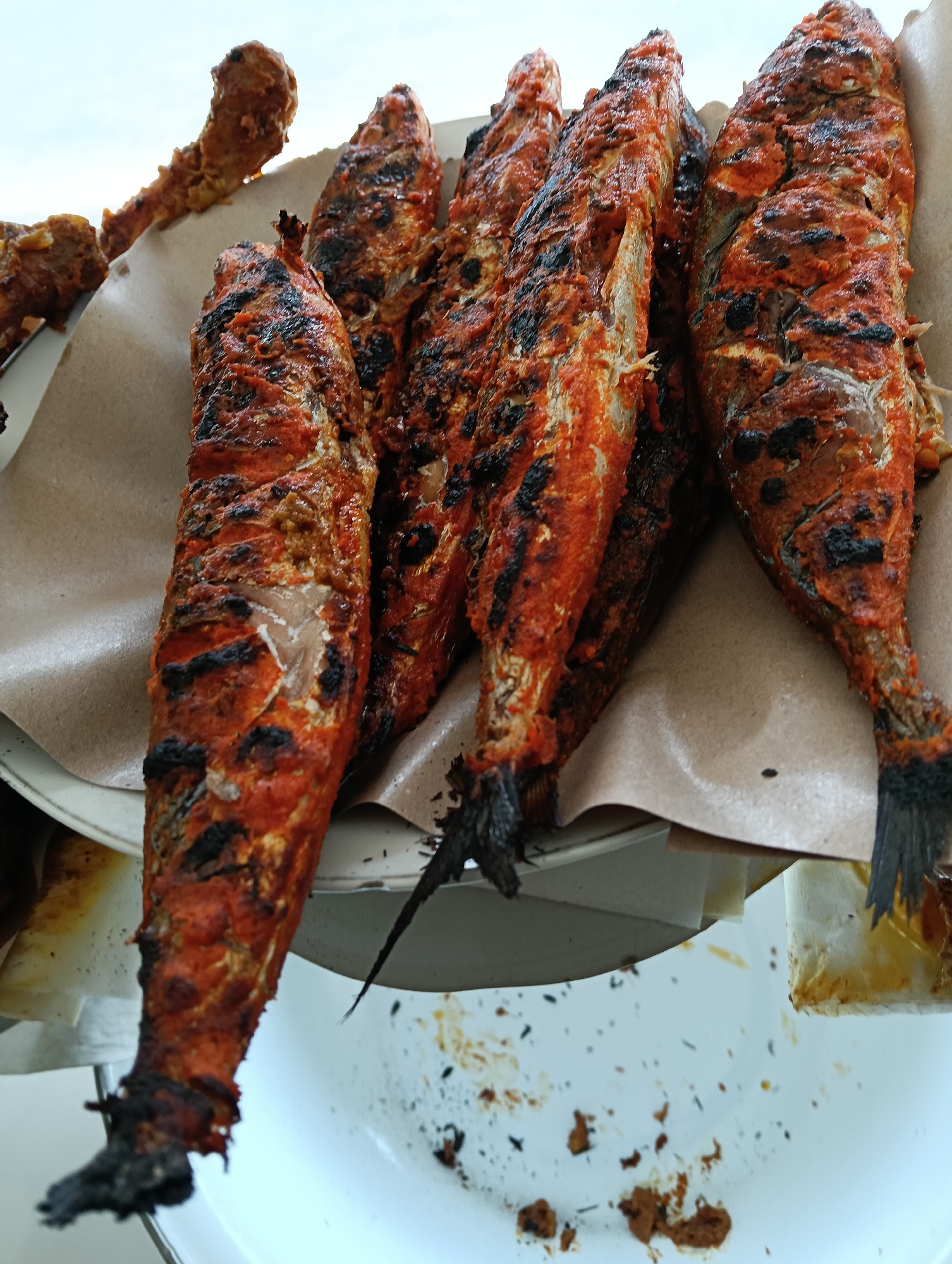 A close-up, slightly overhead shot captures a platter of grilled fish seasoned with a red paste and lightly charred. The fish, appearing to be whole and of a moderate size, are arranged on a light brown, textured paper liner atop a white plate or serving dish. The seasoning is visibly thick and unevenly distributed, with some areas showing the fish's scales beneath. The lighting suggests an outdoor or well-lit indoor setting, with a bright, overcast sky visible in the upper portion of the frame. The background is blurred, but hints of other food items and servingware are present. The scene is devoid of any human presence and evokes a sense of simple, rustic food preparation. The image is presented without any contextual information regarding its specific location in Banjarnegoro, Indonesia, or any anecdotal details.