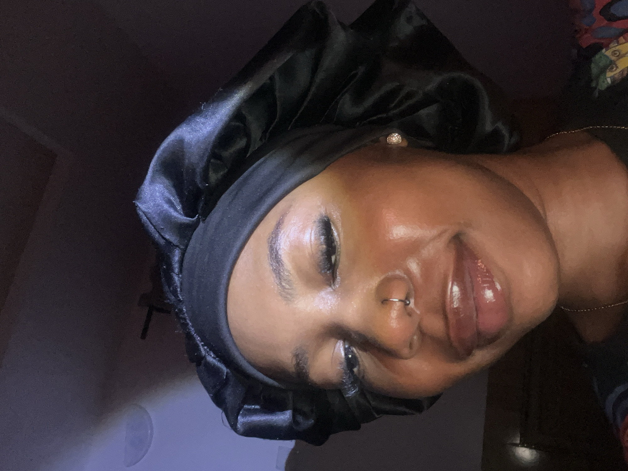 A close-up, selfie-style portrait of a young Black woman wearing a black satin bonnet and a gold nose ring. She has on lip gloss and her eyelashes appear to be falsies. She is looking downwards and slightly to her right, with a subtle smile. A diamond stud earring is visible on her right ear. The background is dark, with a faint blue light illuminating the upper left corner of the image, suggesting an indoor setting at night, potentially in a bedroom or dimly lit room in Rafin Par, Nigeria.