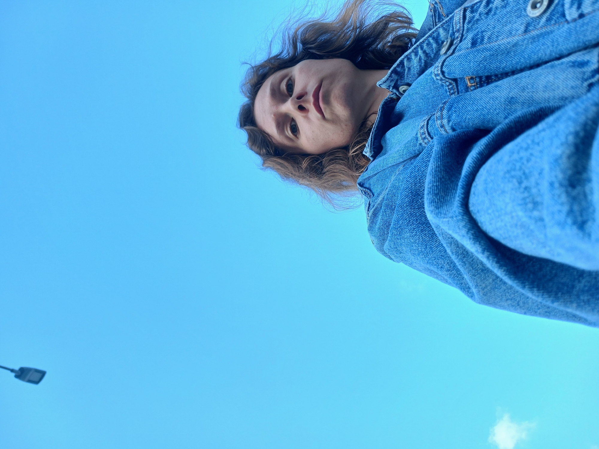 A low-angle, wide shot shows a person wearing a denim jacket against a bright blue sky. The person is looking down towards the camera. A street light is visible in the upper left corner of the frame. The image appears to be taken outdoors in an urban setting, possibly Yekaterinburg, Russia, given the context.