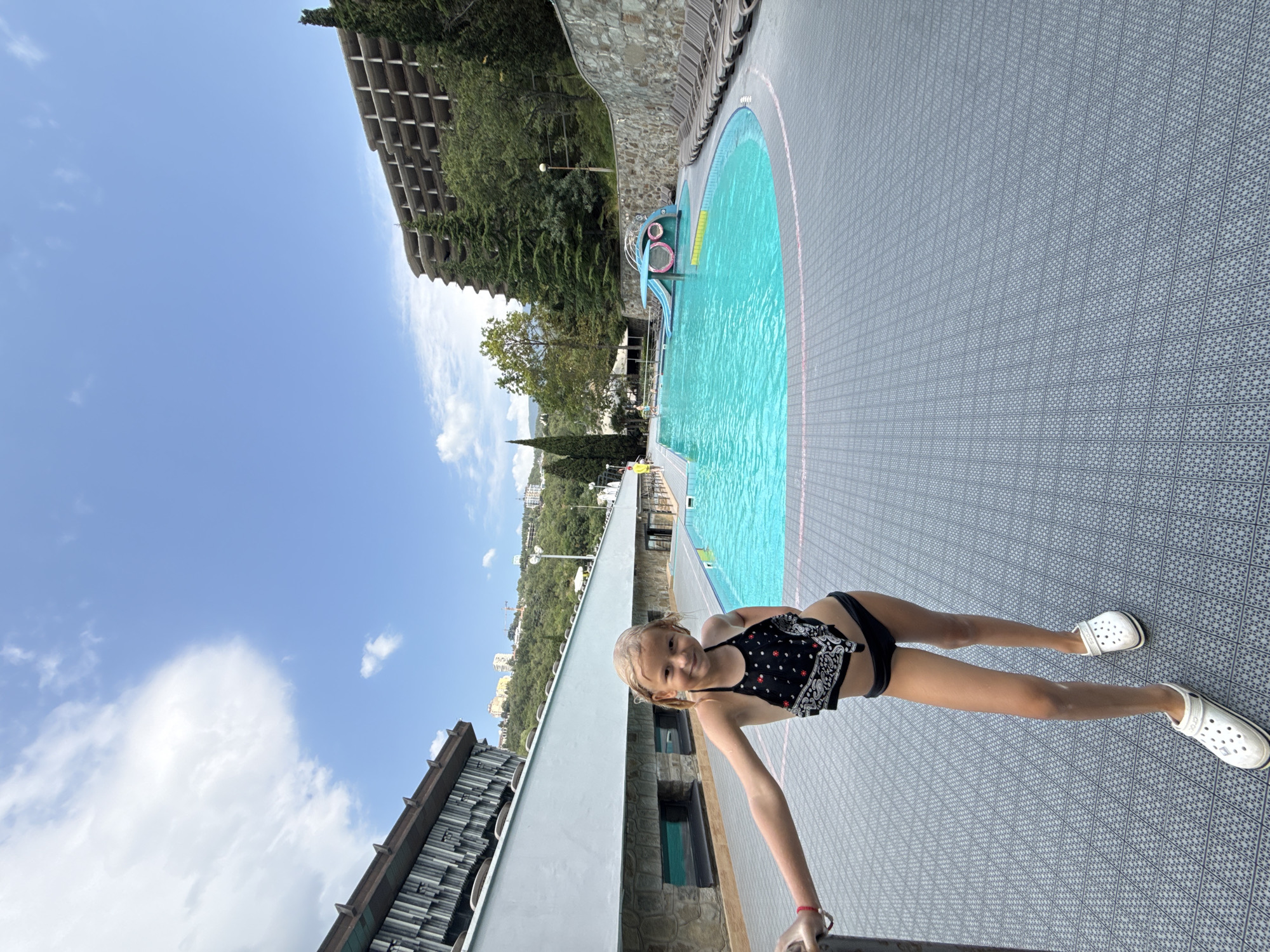 A young girl with blonde hair, wearing a black and white patterned bikini and white Crocs, poses poolside. She is leaning on a railing with her right arm. The setting is an outdoor swimming pool with turquoise water and a grey tiled deck. In the background, a large, multi-story building with a curved facade is visible, covered in greenery, and a stone wall with a balustrade runs along the edge of the pool. The sky is a clear blue with some scattered white clouds, suggesting it is daytime and the weather is pleasant. The girl has a slight smile and appears relaxed. The location appears to be a resort or hotel in a hilly, possibly coastal, area, given the vegetation and distant buildings.