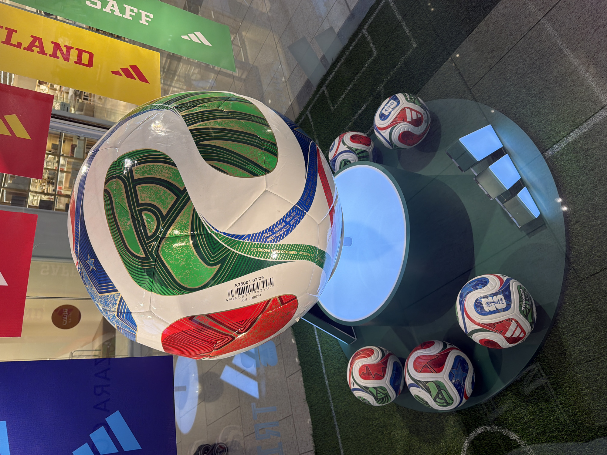 An Adidas Brazuca World Cup official match ball is displayed on a light-up pedestal in front of a store window. Smaller Adidas balls are placed around the pedestal on a green turf display with white soccer field markings. Vertical banners with the Adidas logo and text are visible behind the display. The scene appears to be an indoor retail environment.