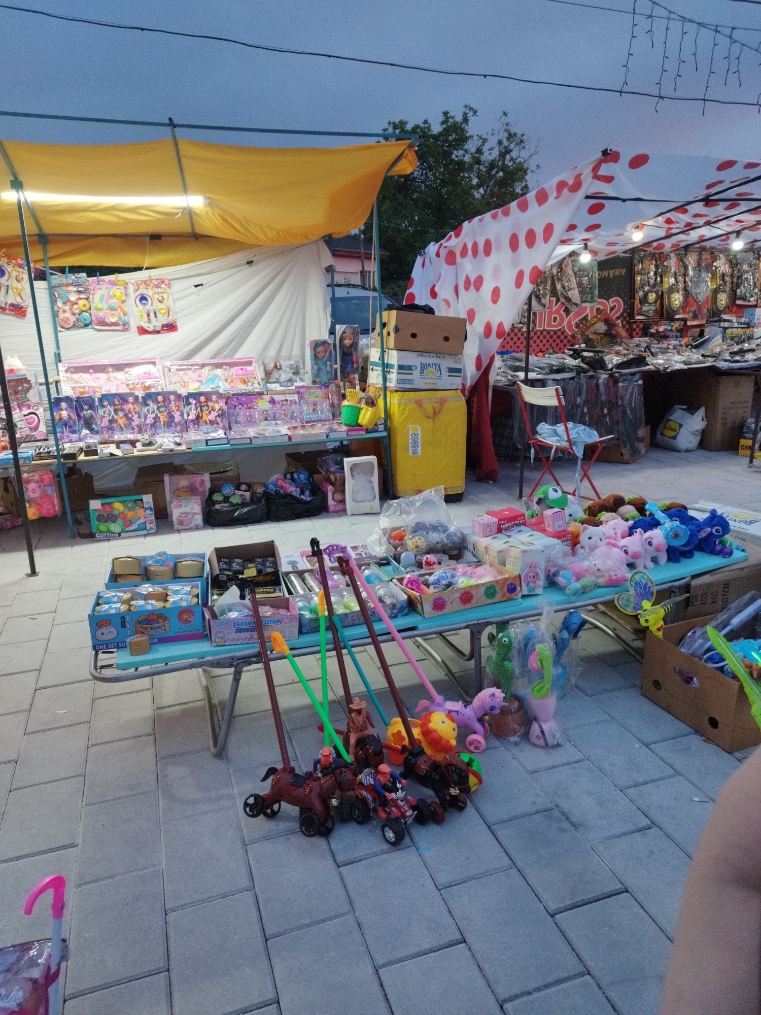 This image shows an outdoor marketplace in an unidentified city. The scene is cluttered with various toys and merchandise for sale, spread across tables under two different tents. One tent has a yellow canopy, and the other has a white canopy with red polka dots. The goods range from stuffed animals and dolls to toy vehicles and various boxed toys. A stack of cardboard boxes labeled "BONITA" stands near the red polka-dot tent. The ground is paved with gray bricks.