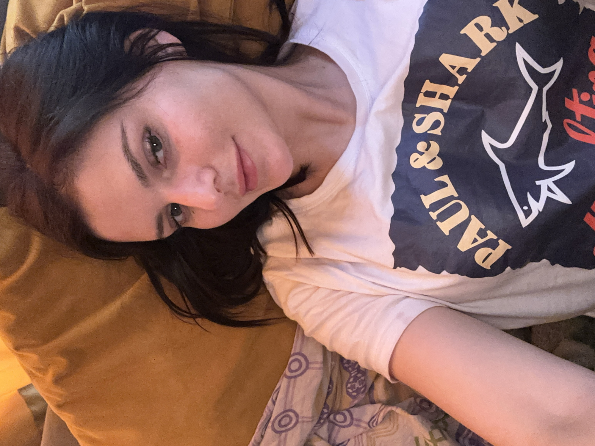 A young woman with dark hair and brown eyes is lying down, looking towards the camera. She is wearing a white t-shirt with a navy blue graphic that features the words "PAUL & SHARK" and a shark illustration. The woman is against a golden-colored cushion or pillow. To her left, a part of a patterned blanket is visible. The image appears to be taken indoors, possibly in a home setting. There are no discernible geographical markers to indicate a specific city or location.