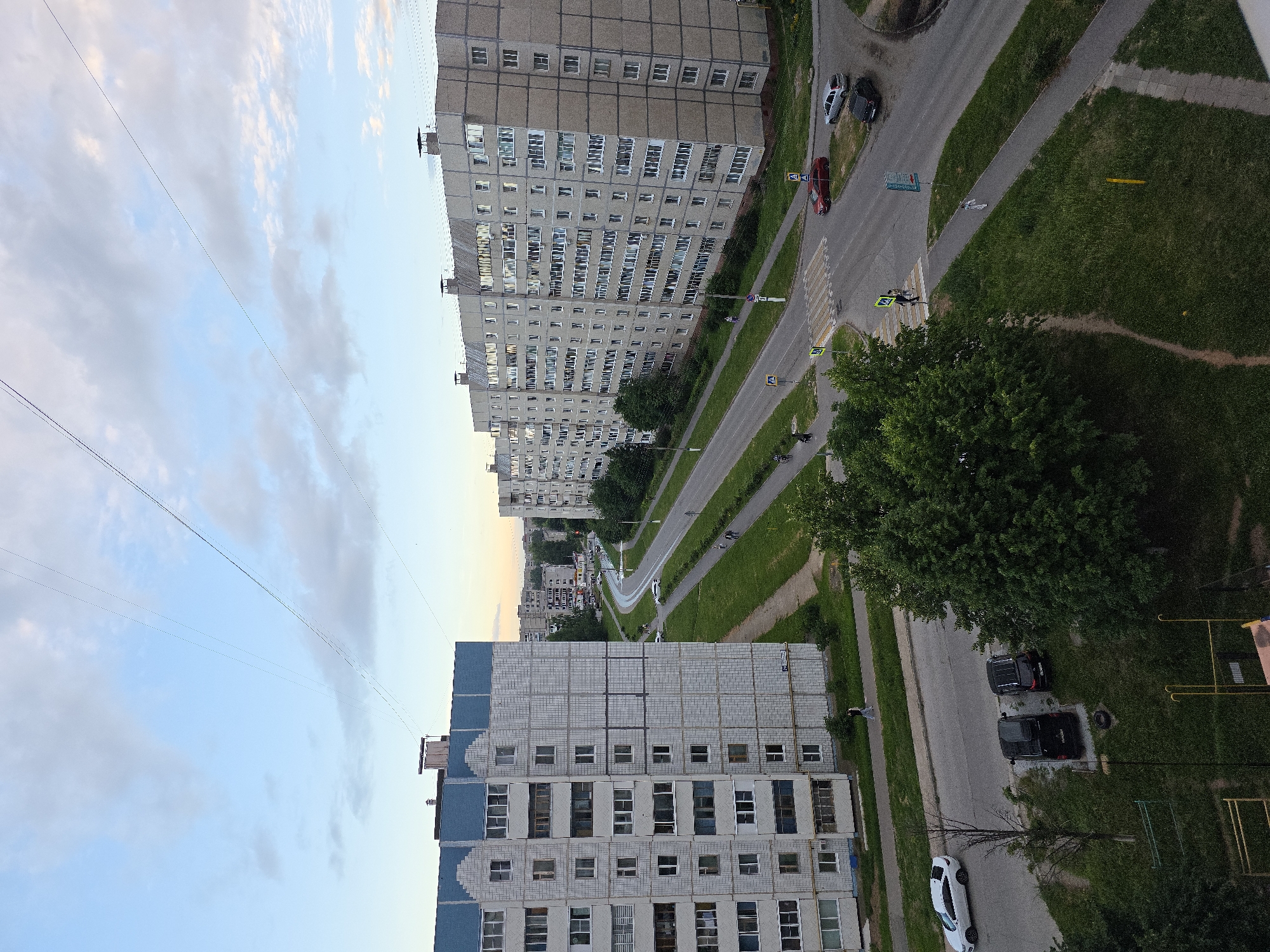 The image depicts a street scene in Novocheboksarsk, Russia, with two large, multi-story apartment buildings dominating the foreground and middle ground. The building on the left is lighter in color with blue accents on its upper floors and features numerous windows. The building on the right is a long, grey structure with a multitude of windows. A road runs between the buildings, with traffic and pedestrian activity visible. Several cars are parked or moving on the road, including a white sedan, a silver sedan, a black SUV, and a red car. Pedestrians can be seen walking on sidewalks and crossing the street at a crosswalk. Green grass and trees are present, indicating a residential area with some landscaping. The sky is partly cloudy, with hints of twilight. Overhead, power lines are strung across the sky. The overall impression is that of a typical urban residential neighborhood in the late afternoon or early evening.