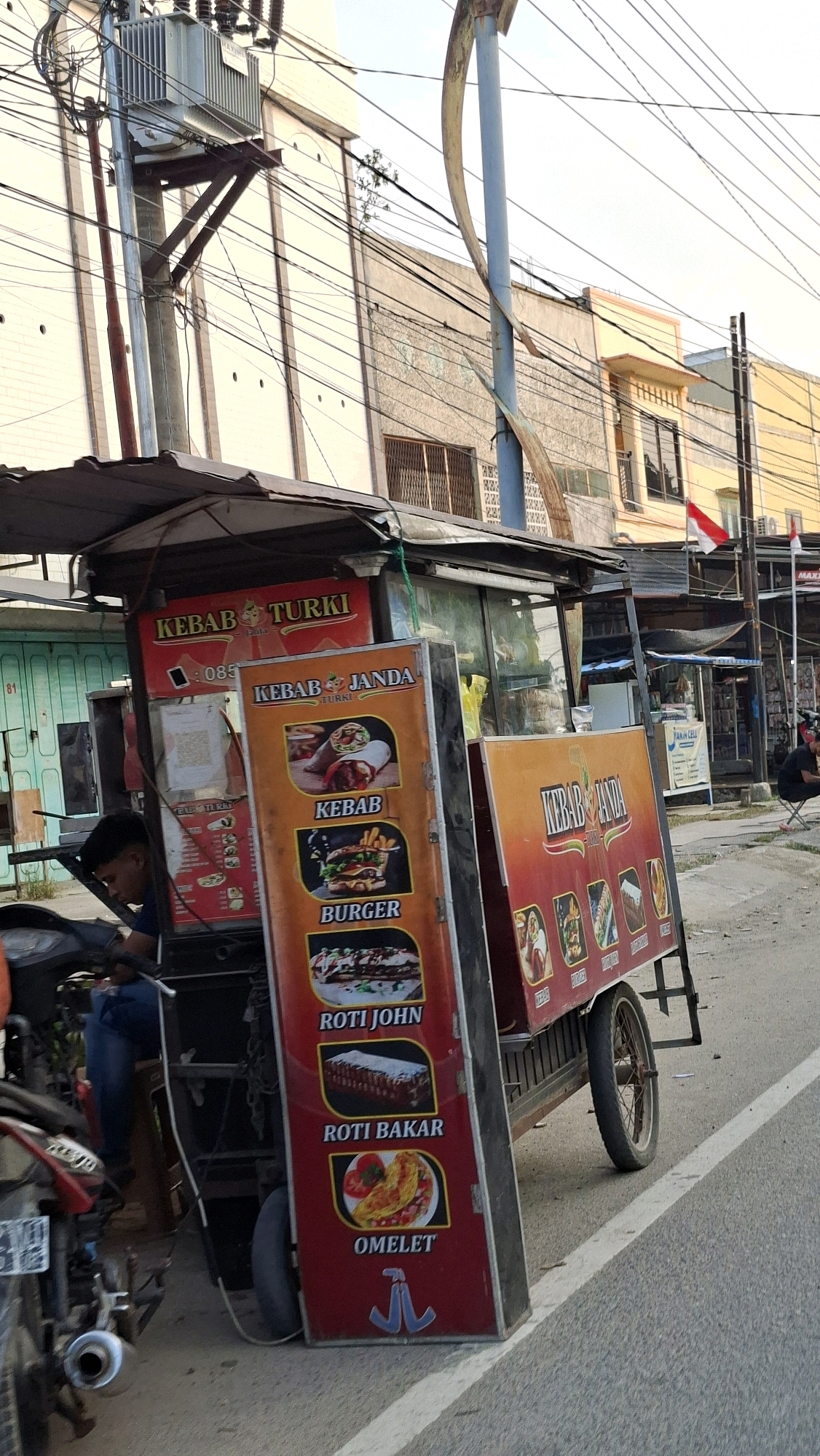The image captures a street scene in Lhokseumawe, Indonesia, focusing on a food cart vendor. The cart is adorned with colorful signs advertising various food items such as "Kebab Turki," "Kebab Janda," "Burger," "Roti John," "Roti Bakar," and "Omelet." A young man, presumably the vendor or an assistant, is seated on a motorcycle next to the cart, looking towards the left. The cart is parked on the side of a paved road, with buildings lining the street in the background. The buildings appear to be a mix of commercial establishments, with some displaying signs and awnings. The sky above is overcast, suggesting it might be late afternoon or early morning. The overall atmosphere is that of a bustling urban environment, with a focus on local street food culture.
