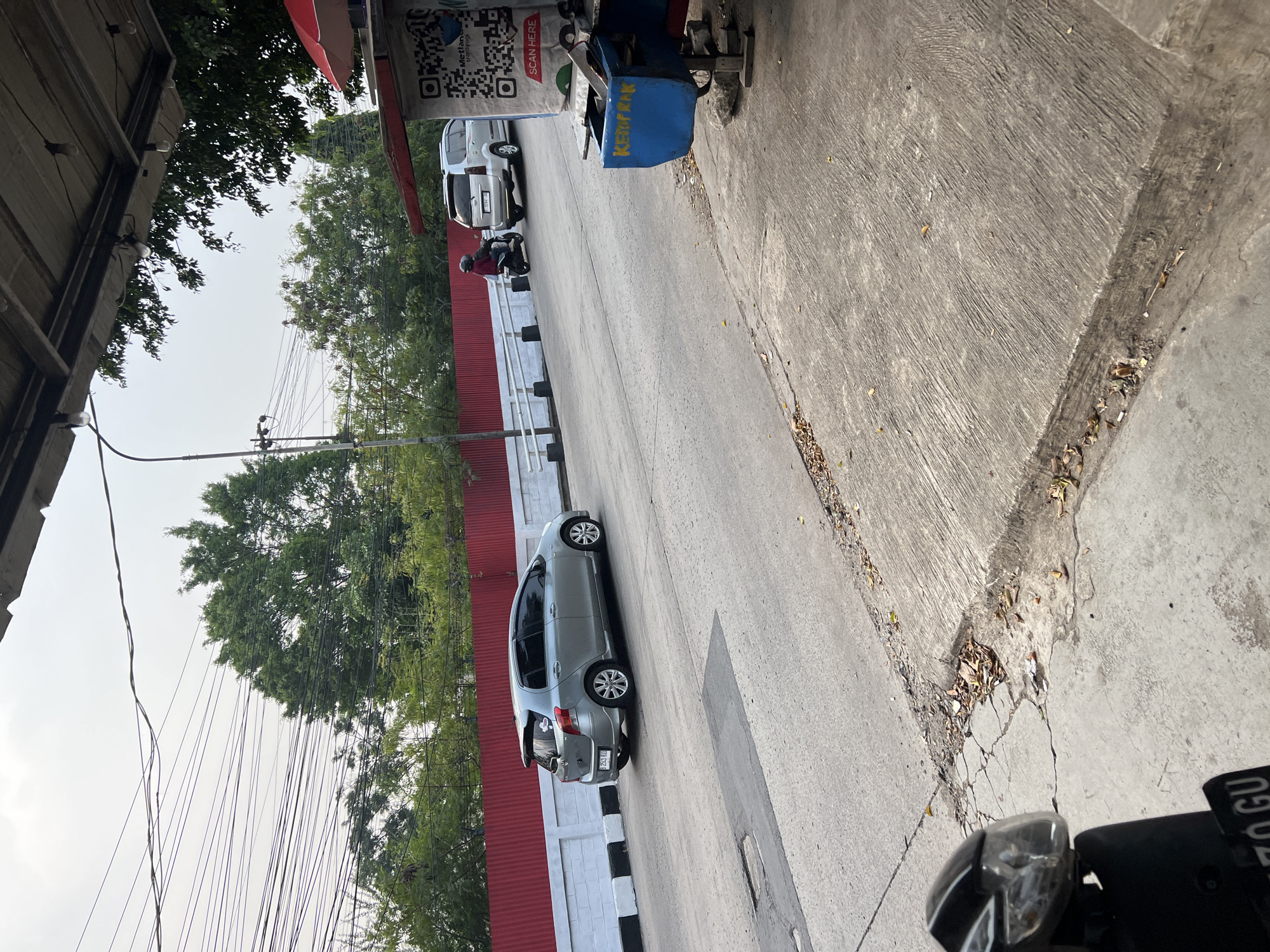The image shows a street scene in Citeureup, Indonesia, with a silver car driving on the road and a motorcycle with a rider in the distance. There is a red fence along the side of the road, and trees are visible behind the fence. In the foreground, a portion of a motorcycle is visible on the left side of the image. On the right side, there is a food cart with a QR code and the text "SCAN HERE" and "KETOPRAK" visible. The sky is overcast.