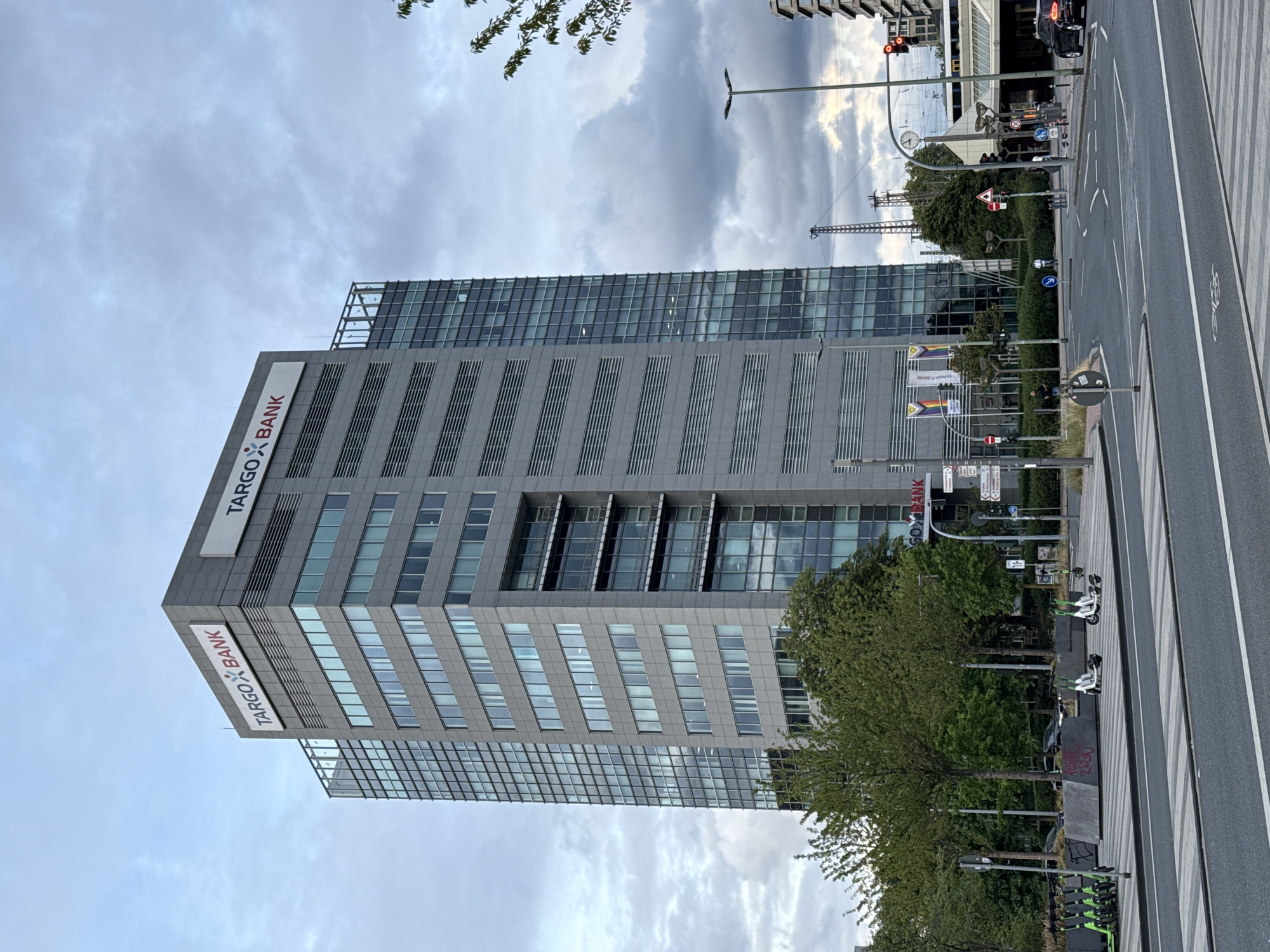 A medium shot captures a tall, modern office building in Duisburg, Germany, on a cloudy day. The building, emblazoned with "TARG X BANK" signage, is a prominent feature, with its grey facade and rows of windows. In the foreground, a street with pedestrian crossings and traffic lanes is visible. Several electric scooters are parked along the sidewalk, and a few trees provide a touch of greenery. In the background, other buildings, streetlights, and what appear to be train tracks are present, indicating an urban environment. The sky is filled with dramatic, grey clouds, suggesting the possibility of rain.