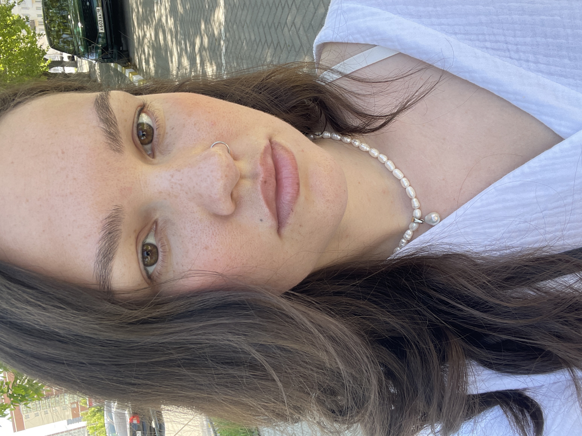 A close-up selfie captures a woman with brown hair and hazel eyes wearing a white top and a pearl necklace. She has fair skin with freckles and a nose ring. The background shows glimpses of a city environment, including parts of a building, trees, and vehicles, potentially located in Izhevsk, Russia. A parked black car with a Russian license plate is visible in the background. The scene appears to be outdoors during daylight.