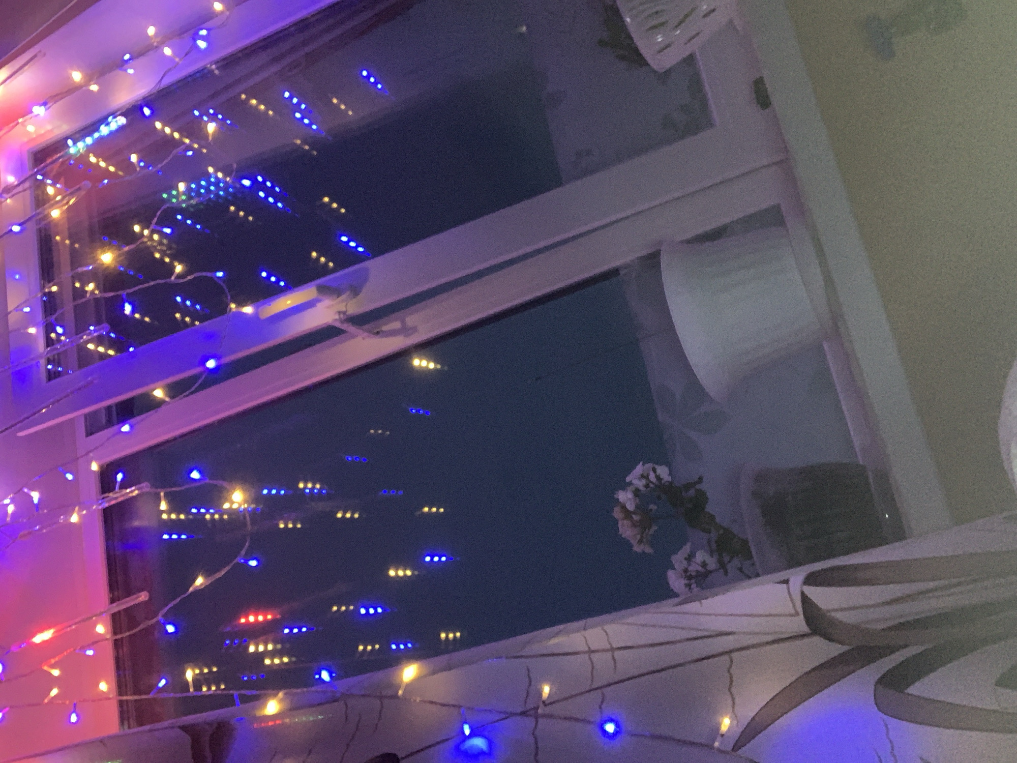 This media file captures a close-up view of a window decorated with colorful LED string lights, creating a festive and cozy atmosphere. The lights, predominantly blue and yellow, with some red and pink hues, are draped along the window frame and appear to be reflected in the glass. The scene suggests an indoor setting, likely a home, with the window looking out onto a dark exterior, implying it is nighttime or dusk.
In the reflection on the window, a faint pattern of lights can be seen, possibly from other buildings or streetlights in Leninogorsk, Russia. A few sparse, white flowers on a branch are visible in the upper right portion of the reflection, adding a delicate natural element to the otherwise artificial display of lights. The lighting is warm and inviting, with the bright LED lights casting colorful glows on the surrounding surfaces. The overall mood conveyed is one of cheerfulness and tranquility, characteristic of holiday decorations or a personal touch to an living space. No visible text is present in the image.