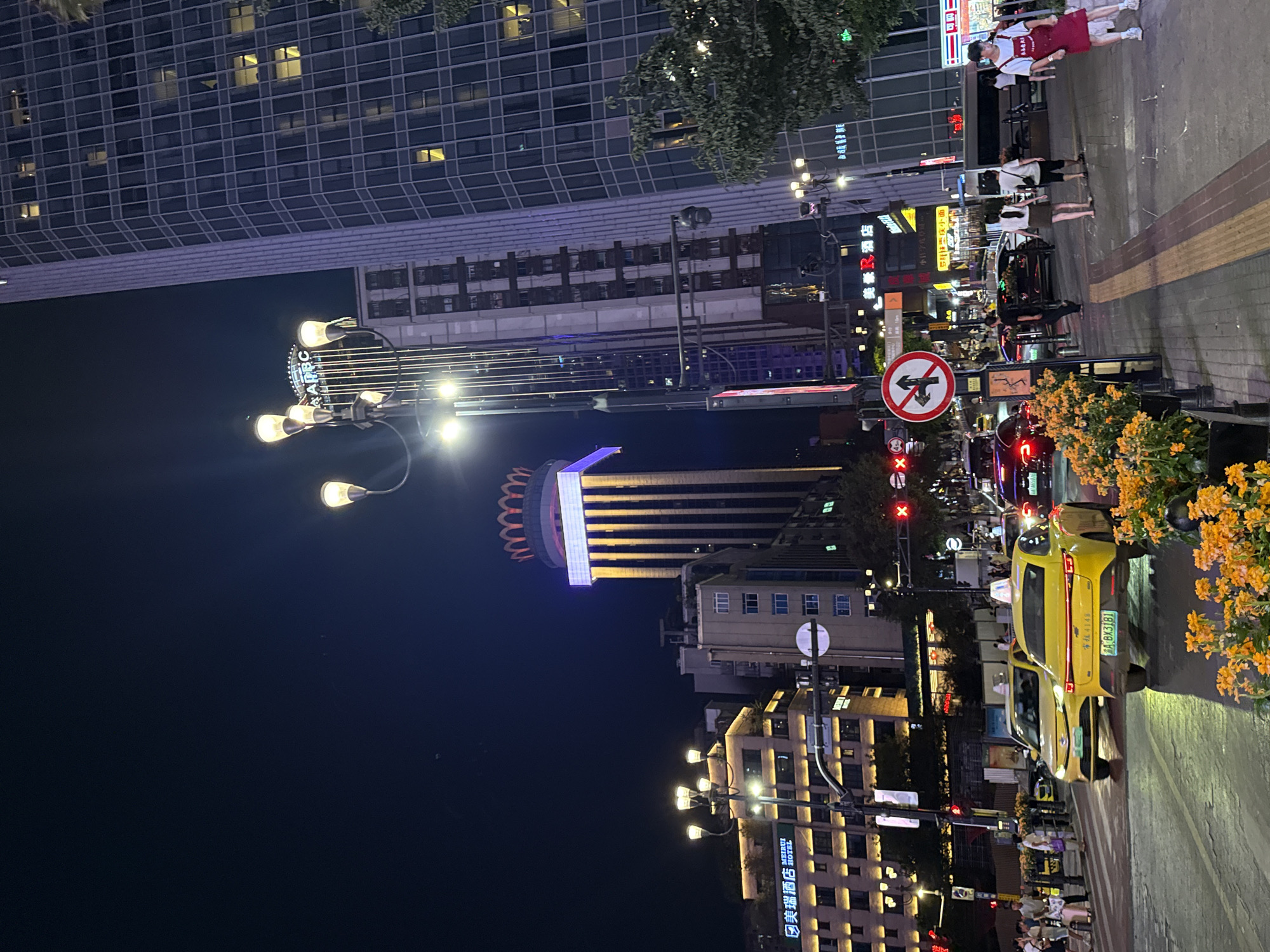 This nighttime photograph captures a bustling street scene in Yuzhong District, China. The image is oriented vertically, presenting a view looking up from street level. Towering buildings with illuminated windows dominate the background, suggesting an urban environment. In the foreground, a bright yellow taxi is prominently featured, its license plate partially visible as "A BX3181" and a partial Chinese license plate. The taxi is positioned on a paved road with a pedestrian crossing.
Several people are visible on the sidewalk, some walking and others standing. They appear to be casually dressed, contributing to the everyday atmosphere of the scene. The presence of streetlights and illuminated signs indicates that it is evening or night. One sign displays the text "美瑞酒店" (Meirui Hotel) and another, further down, shows "美豪酒店" (Meihao Hotel), suggesting the area is a commercial hub with accommodation options.
Other visible text includes "APEC" on a lighted sign and what appears to be Chinese characters on various storefronts and advertisements. A traffic sign with a red circle and a diagonal line, indicating a prohibition, is also present. The overall impression is that of a lively urban setting at night, with vehicles, pedestrians, and illuminated architecture creating a dynamic and vibrant scene. The lighting from the buildings and signs casts a warm glow on the street.