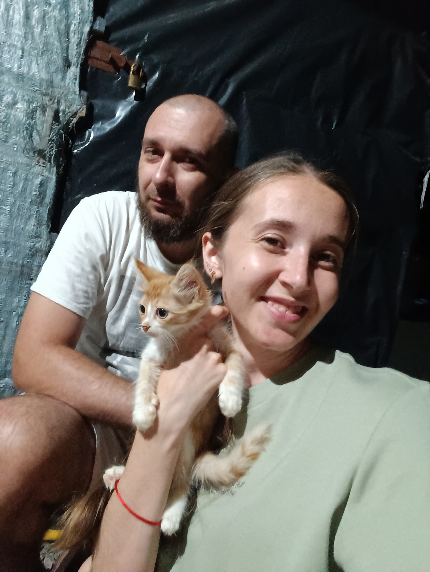 The image shows a selfie of a man and a woman holding a ginger kitten. The man, with a bald head and beard, is wearing a white t-shirt. The woman, smiling broadly, is wearing a light green t-shirt and has a red bracelet on her wrist. The kitten is held by the woman and is looking to the side. The background appears to be a dimly lit, possibly makeshift interior space, with a black tarp-like material and a textured, possibly corrugated metal wall. A rusty padlock is visible on the wall in the background. The location is Vypasne, Ukraine.
