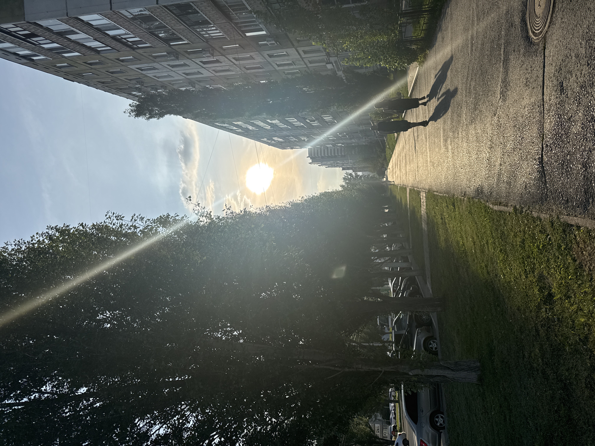 The image depicts a sun-drenched street scene in Ufa, Russia. Two individuals, appearing to be a couple, are walking away from the viewer down a paved sidewalk. They are casting long shadows on the glistening asphalt, indicating it is likely late afternoon or early evening. The sun is bright and positioned high in the sky, with rays of light streaming diagonally across the frame, creating lens flare effects.
To the left of the sidewalk, a grassy area is lined with parked cars. Further left, a row of trees with lush green foliage creates a natural border. To the right of the sidewalk, a multi-story apartment building with numerous windows and visible air conditioning units dominates the view. The building's facade is a neutral color, likely concrete or stucco, and shows signs of age. In the distance, more apartment buildings and trees are visible, suggesting an urban or suburban residential area. The overall atmosphere is one of a warm, sunny day in a populated area.