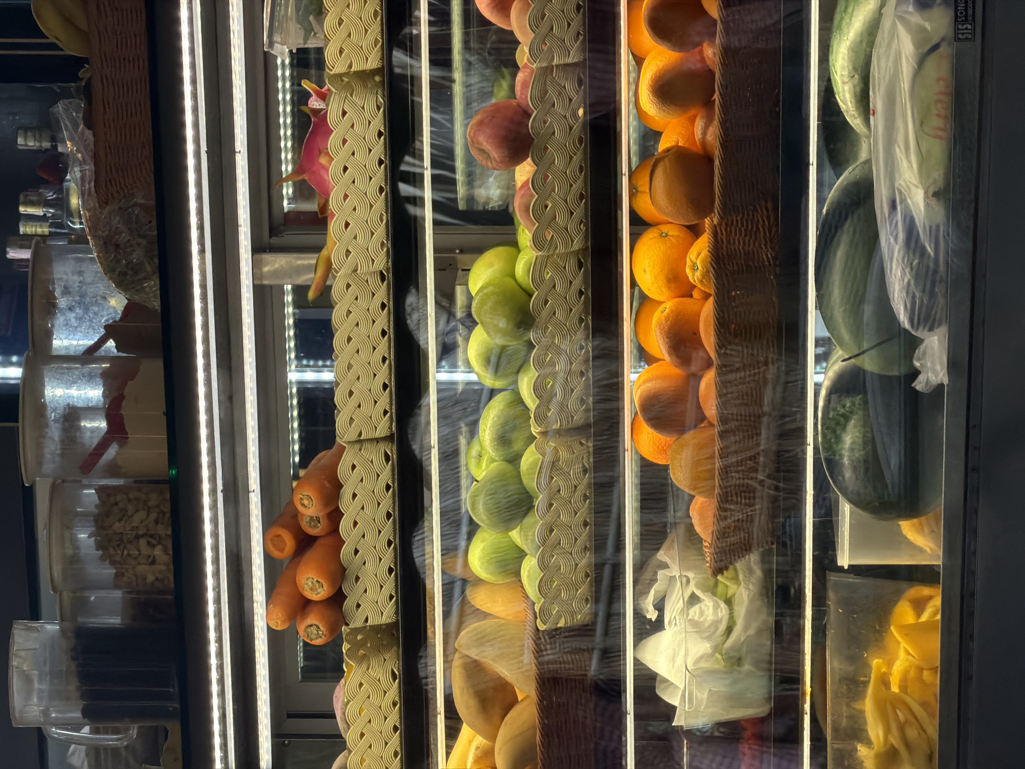 A produce cooler with vertical shelves is stocked with fruits and vegetables, including bananas, dragon fruit, apples, green apples, oranges, watermelons, mangoes, and carrots. These items are arranged neatly in their respective sections. Some items, like watermelons, are wrapped in clear plastic. The cooler has a built-in light to showcase the produce. The location is in Choeng Thale, Thailand.