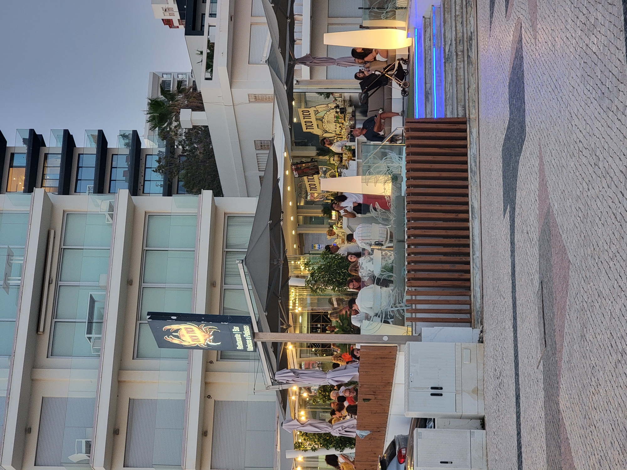 This is a medium shot of a restaurant called "Tico Tico" located in Quarteira, Portugal, at dusk. The restaurant has an outdoor seating area with patrons dining under umbrellas. The building behind the restaurant is modern with balconies. In the foreground, a man is sitting on a sofa with a baby stroller next to him. The pavement is made of cobblestones, and there are large, illuminated pillars on either side of a staircase with blue lighting. The sky is overcast and a pale blue.
