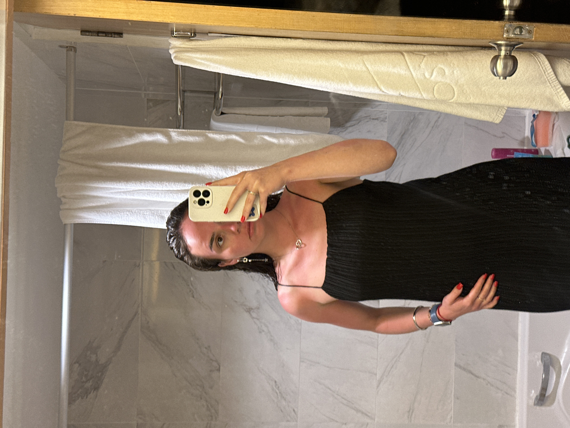 A woman stands in a bathroom taking a selfie. She is wearing a black spaghetti strap dress and has red nail polish on her fingers. Her hair is dark, and she wears a necklace with a heart-shaped pendant. A smartwatch adorns her left wrist. The bathroom features white marble tile on the walls and floor. A white towel hangs above a towel rack. Toothpaste and other toiletries are visible in the background. The bathroom appears to be in a hotel room in Pattaya City, Thailand.