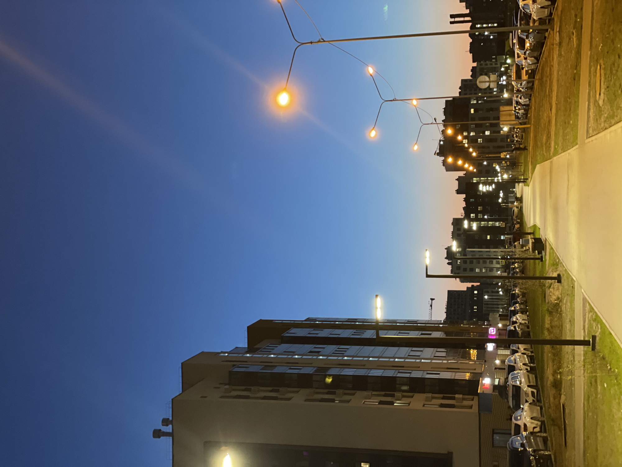 This is a nighttime image taken in the city of Kazan, Russia. The photo is oriented with the sky at the top and ground at the bottom.
The sky transitions from a deep blue in the lower left to a lighter blue and orange near the horizon at the top of the frame. Streaks of light are visible in the upper left sky.
The scene depicts a residential area with apartment buildings. The buildings are illuminated by streetlights and lights from within the apartments. The buildings are modern in design with multiple stories and many windows.
A street or walkway is visible on the right side, illuminated by streetlights. A line of parked cars is visible along the side of the road near the buildings. Streetlights are visible on both sides of the road. A grass area is situated between the road and the buildings.
The scene is set during twilight or early evening, with artificial lighting illuminating the surroundings.