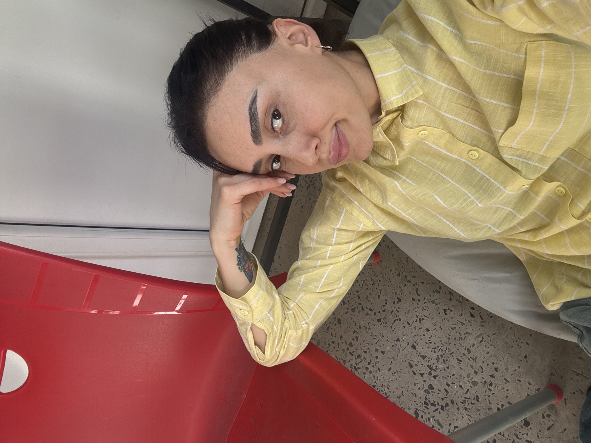 A woman is seated in a red plastic chair with metal legs indoors. She wears a yellow and white plaid shirt and has dark hair pulled back. She has a tattoo on her left forearm. The flooring is a speckled gray pattern. A white wall is visible in the background. The location cannot be determined from the image.