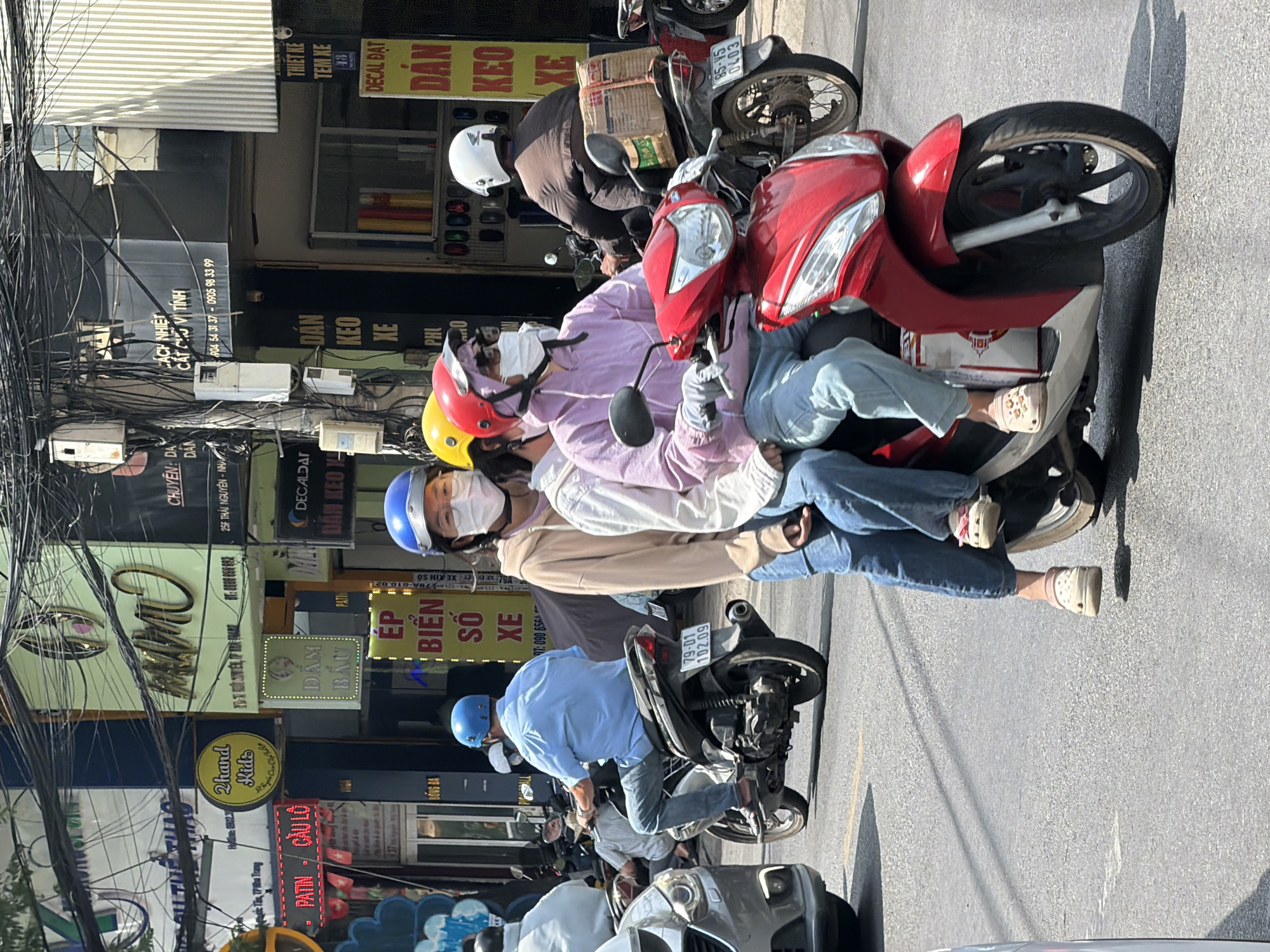 The image depicts a street scene in Nha Trang, Vietnam, featuring a red scooter with three people riding on it. The driver, wearing a blue helmet and a light blue short-sleeved shirt, is a male. The two passengers behind him are females; the one in the middle wears a yellow helmet and a light brown hoodie, and the rear passenger has a pink hoodie and a white helmet. Both passengers are wearing face masks. The scooter is traveling on a paved road, with other vehicles and buildings visible in the background. The buildings have various signs, some in Vietnamese, suggesting commercial establishments. Overhead, a dense network of electrical wires crisscrosses the sky, typical of urban Vietnamese streetscapes. The lighting suggests it is daytime, with shadows cast on the road.