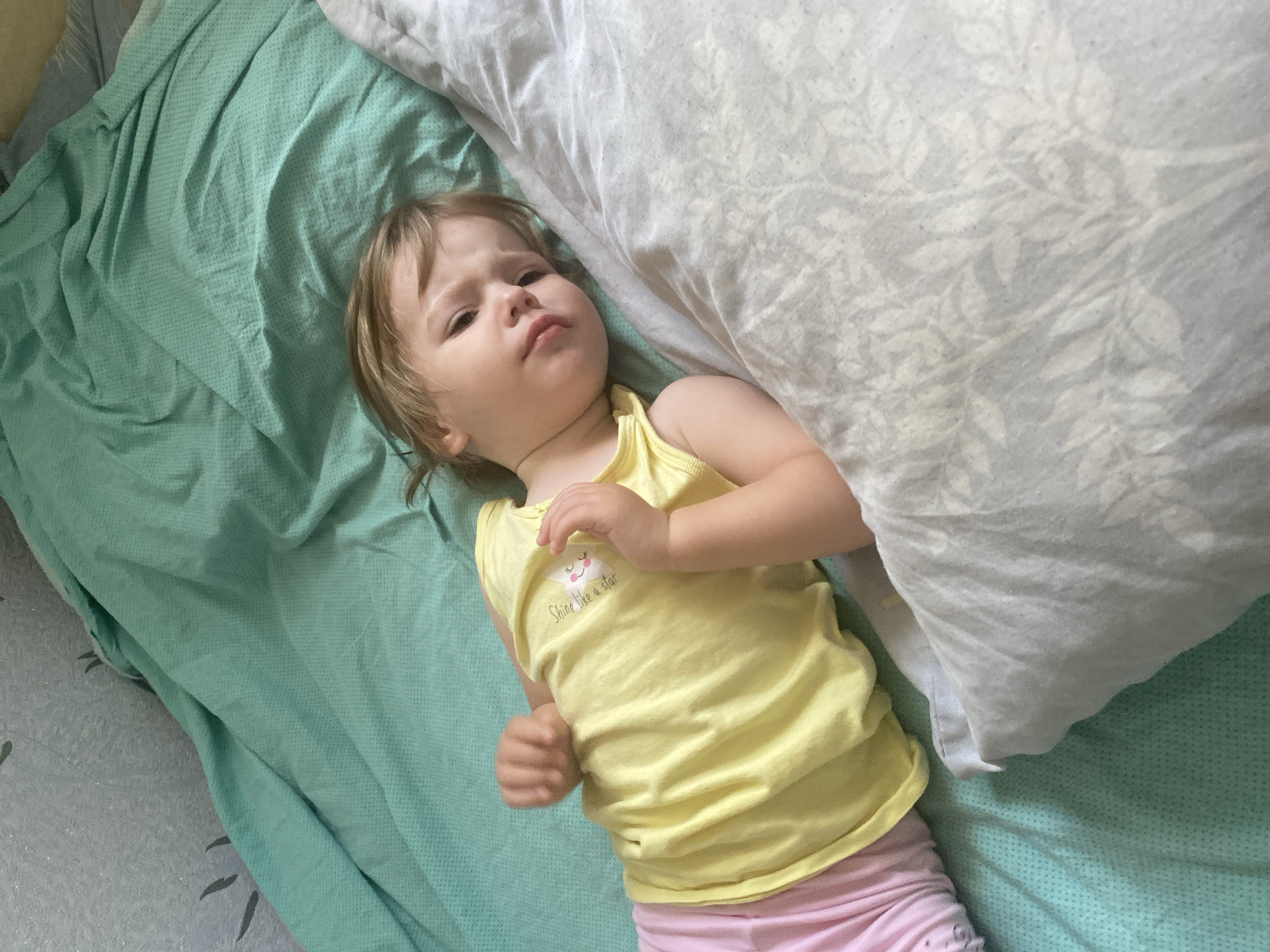 A young child with blonde hair lies on a bed covered by a teal blanket and a patterned gray pillow. The child is wearing a yellow tank top with a graphic of a smiling cloud and star that says "Shine like a star," and pink pants. The child's left arm is bent at the elbow and raised, with their hand near their face. Their right arm is also bent at the elbow, resting on their side. The child's head is turned towards the right, and their gaze is directed slightly upward, with a neutral expression. The lighting suggests it is daytime. The environment appears to be a bedroom.
