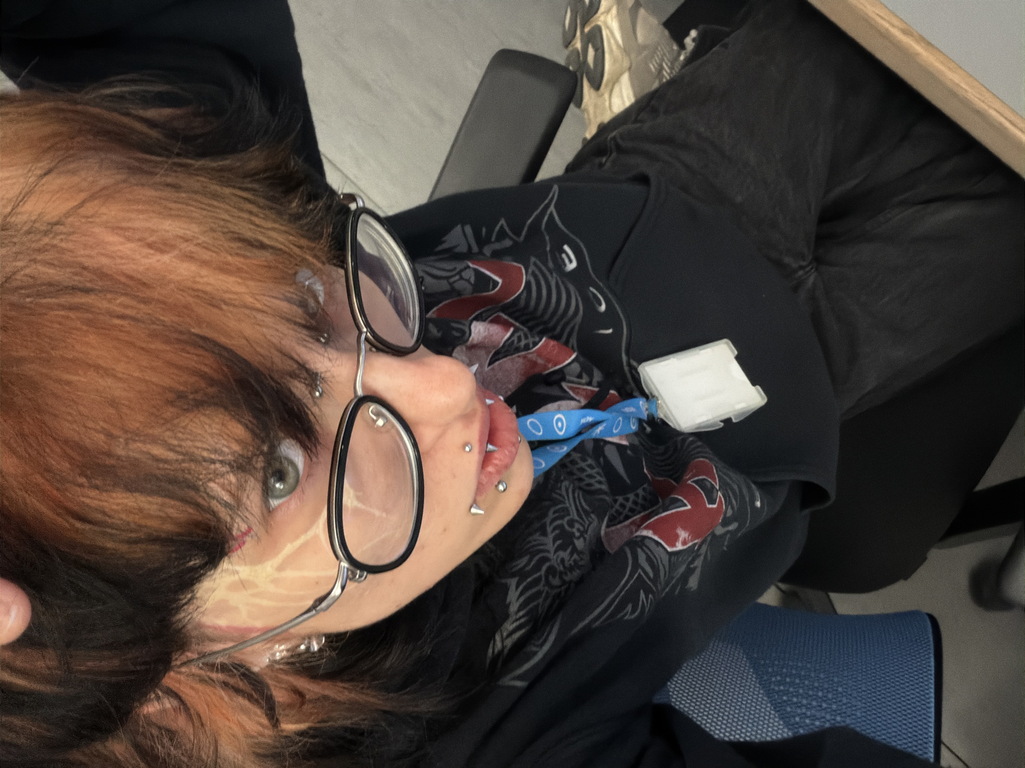 A medium close-up, rotated portrait shows a person with striking two-toned hair, featuring dark roots and reddish-brown mid-lengths and ends. The individual wears round, wire-rimmed glasses and has multiple facial piercings, including a bridge piercing between the eyes, several lip piercings, and ear piercings. Their left eye, a clear green or hazel, looks directly towards the viewer. The left side of their face shows light-colored streaking or marking, possibly intentional artistic makeup or scarification, and a small red mark near the outer corner of the eye.
The person is wearing a dark, long-sleeved top with a grey and red graphic design, possibly depicting a stylized animal or skull, over their chest. Around their neck hangs a blue lanyard with repeating white circular symbols, attached to a white plastic ID card holder.
The setting appears to be indoors, with parts of a dark chair, a wooden desk edge, and a blue mesh chair visible in the background. The lighting suggests an artificial indoor environment. The image is captured from a low angle, with the person looking upwards at the camera, conveying a direct and intense gaze. No distinct text is legible on the lanyard, ID card, or clothing graphic. This scene is located in Busmantsi, Bulgaria.