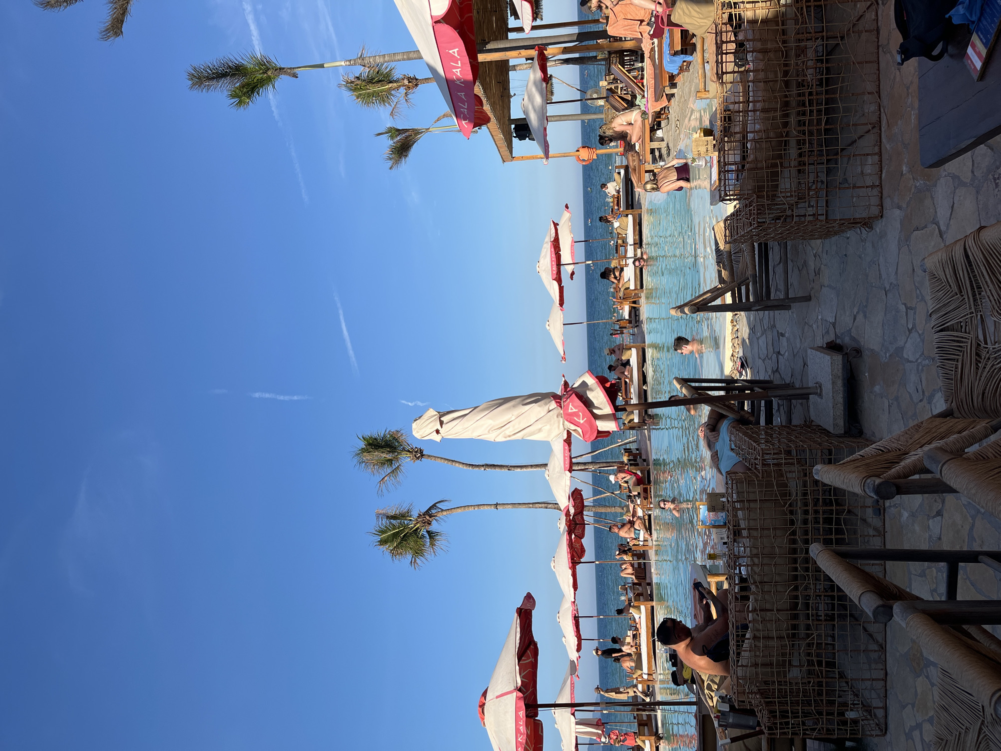 The image shows a beach scene with a clear blue sky and palm trees. There are numerous white and red beach umbrellas shading lounge chairs arranged along the water's edge. People are seen relaxing on the chairs, swimming, and interacting with each other. The area appears to be a popular beach resort or club, evidenced by the beach furniture and sunbathing individuals. The location is not specified in the image, and it cannot be identified as Đà Nẵng, Vietnam, without further information.