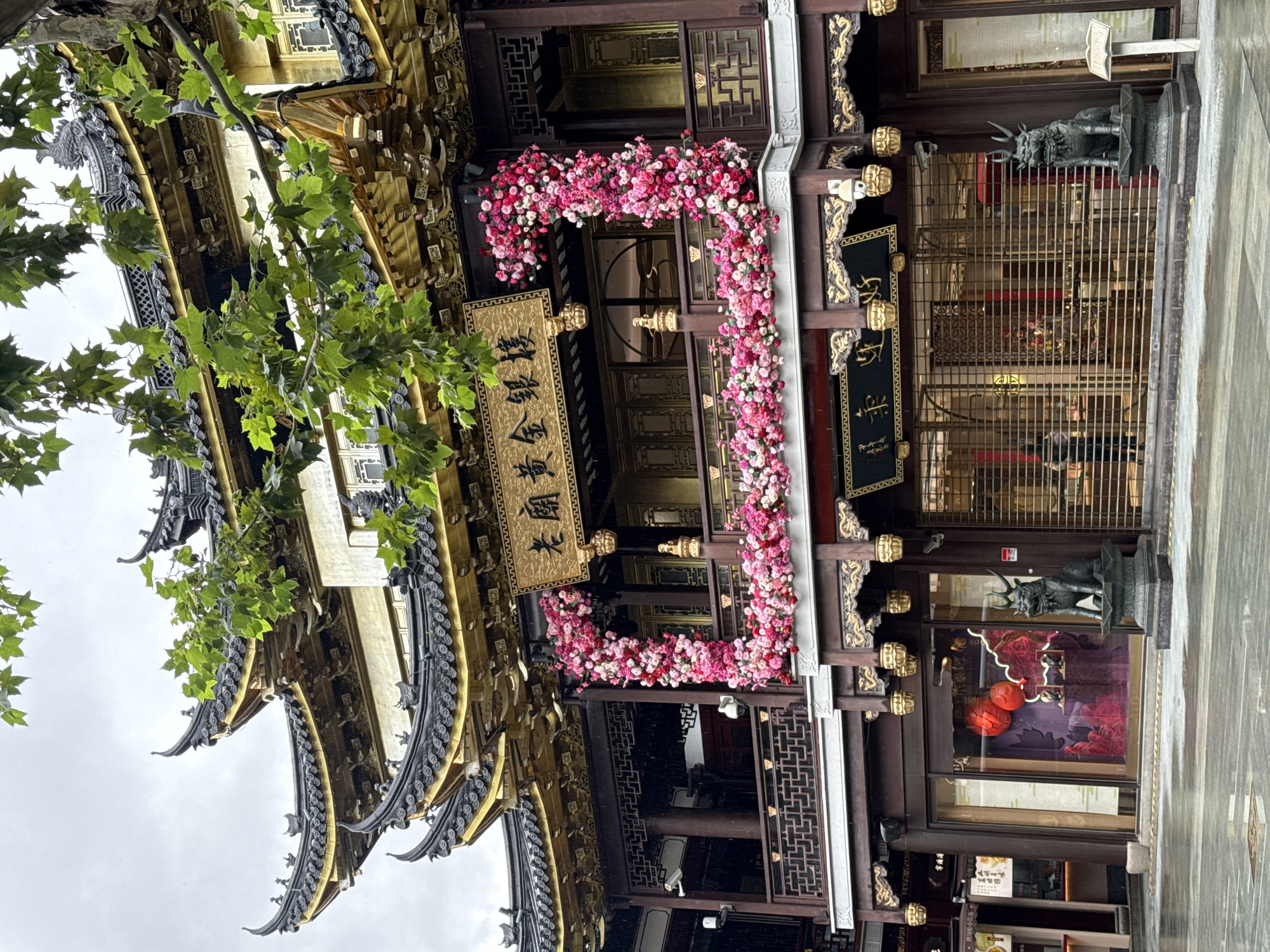 This is a medium shot of the Yu Garden Bazaar in Shanghai, China, on a rainy day. The traditional Chinese architecture is characterized by ornate, multi-tiered roofs with upturned eaves, gilded detailing, and dark wooden structures. A prominent sign with Chinese characters hangs above the entrance. The building is decorated with a large, vibrant floral arrangement of pink and white flowers draped over a balcony. Two decorative stone statues, resembling mythical creatures, flank the entrance on either side of the street. The street in front of the building appears wet and reflective due to the rain, with visible water puddles. The ground is paved with cobblestones. Security cameras are mounted on the building. The lower levels of the building feature shopfronts with closed metal grates, and one shop window displays red lanterns and red floral patterns. A few people are visible inside the shops through the grates. Green foliage from trees hangs down from above the building.