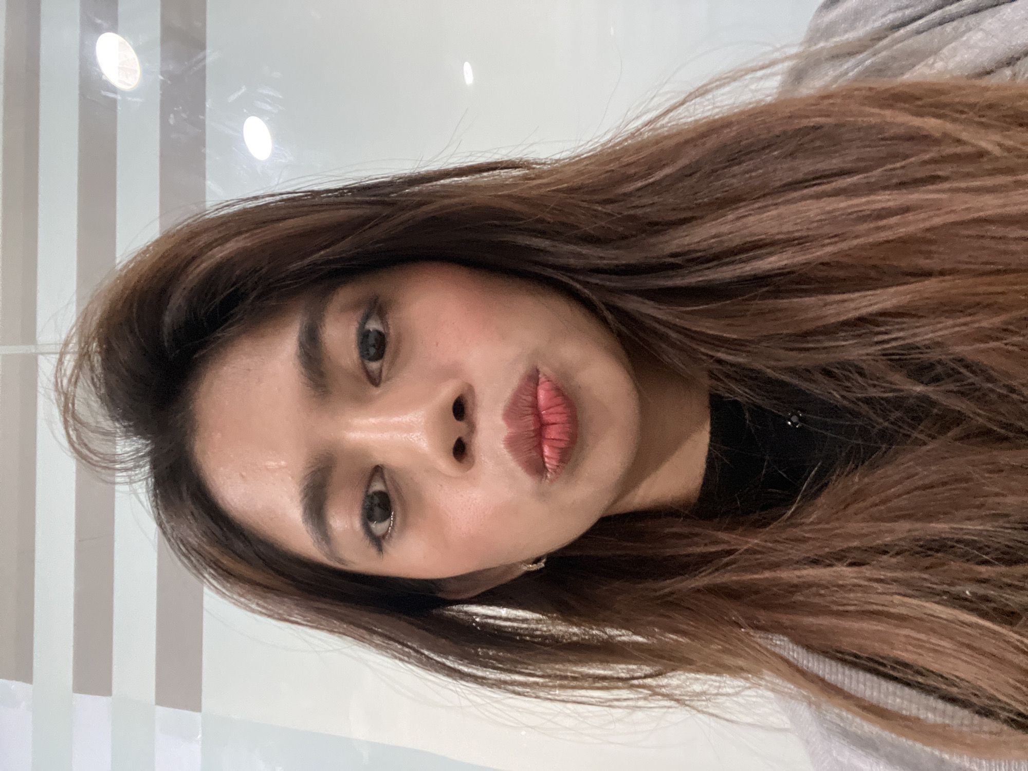 This close-up selfie features a young woman captured indoors, holding her phone vertically, resulting in a horizontally-oriented image. Her long, dark brown hair, with subtle lighter brown tones, frames her face and extends across the frame. She has dark eyes, enhanced with eyeliner and mascara, and her cheeks show a touch of rosy blush. Her lips are pursed in a playful "kiss face" or pouty expression, colored with a pinkish-red lipstick.
She is wearing a dark top, possibly black, partially visible around her neck and shoulder. A light grey or white garment, likely a sweater or jacket, is visible over her right shoulder (viewer's left). The background consists of a light-colored wall with alternating vertical stripes of white/light grey and light brown/beige. The wall surface appears slightly reflective, showing distinct circular reflections of overhead lights. No text is visible in the image. The setting is indoors, with the provided context indicating the location as Carmen, Philippines.