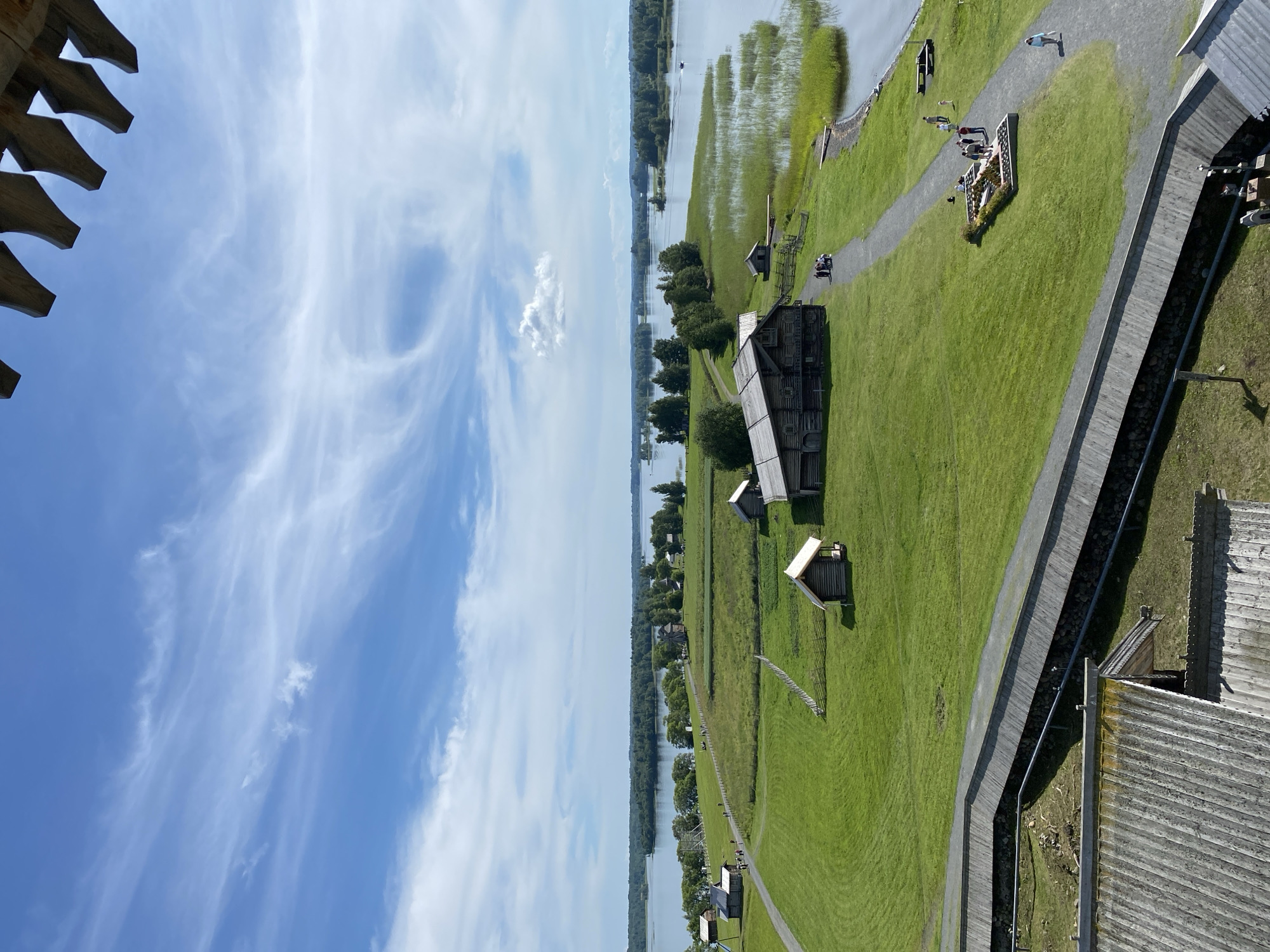 An aerial, eye-level, wide shot captures a historic village with wooden structures and a vast body of water under a bright, partly cloudy sky. The scene is located in Boyarshchina, Russia. In the foreground, a wooden walkway with a railing is visible, leading the eye into the expansive green landscape. Scattered across the grassy fields are several traditional wooden buildings, some appearing to be houses or barns, with thatched or wooden roofs. A larger, prominent wooden structure with a dark roof and visible windows stands towards the center-left of the frame. A gravel path winds through the village, and several small groups of people are seen walking along it, appearing to be tourists. The path leads towards the water's edge, where reeds grow thick along the shore. The calm water stretches out to a horizon dotted with distant, tree-covered islands. The sky above is a vibrant blue, with wispy white clouds creating texture and depth. The overall impression is of a serene, preserved historical site surrounded by natural beauty.